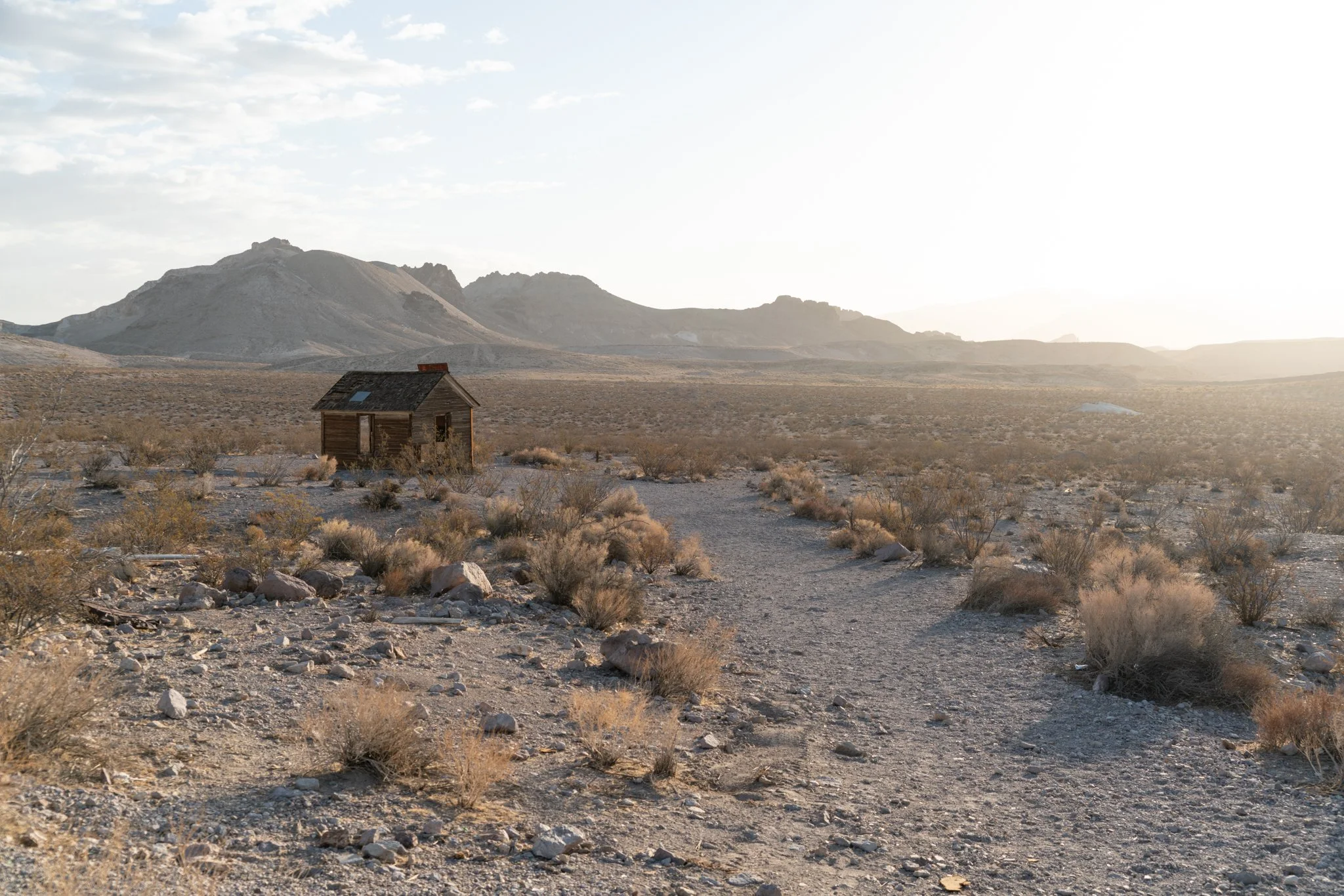 Dust and Distance: The Resonance of Time, limited edition fine art photograph by Jason Sponseller, featuring a weathered desert cabin in Death Valley surrounded by dry brush, distant mountains, pale light, and open desert silence
