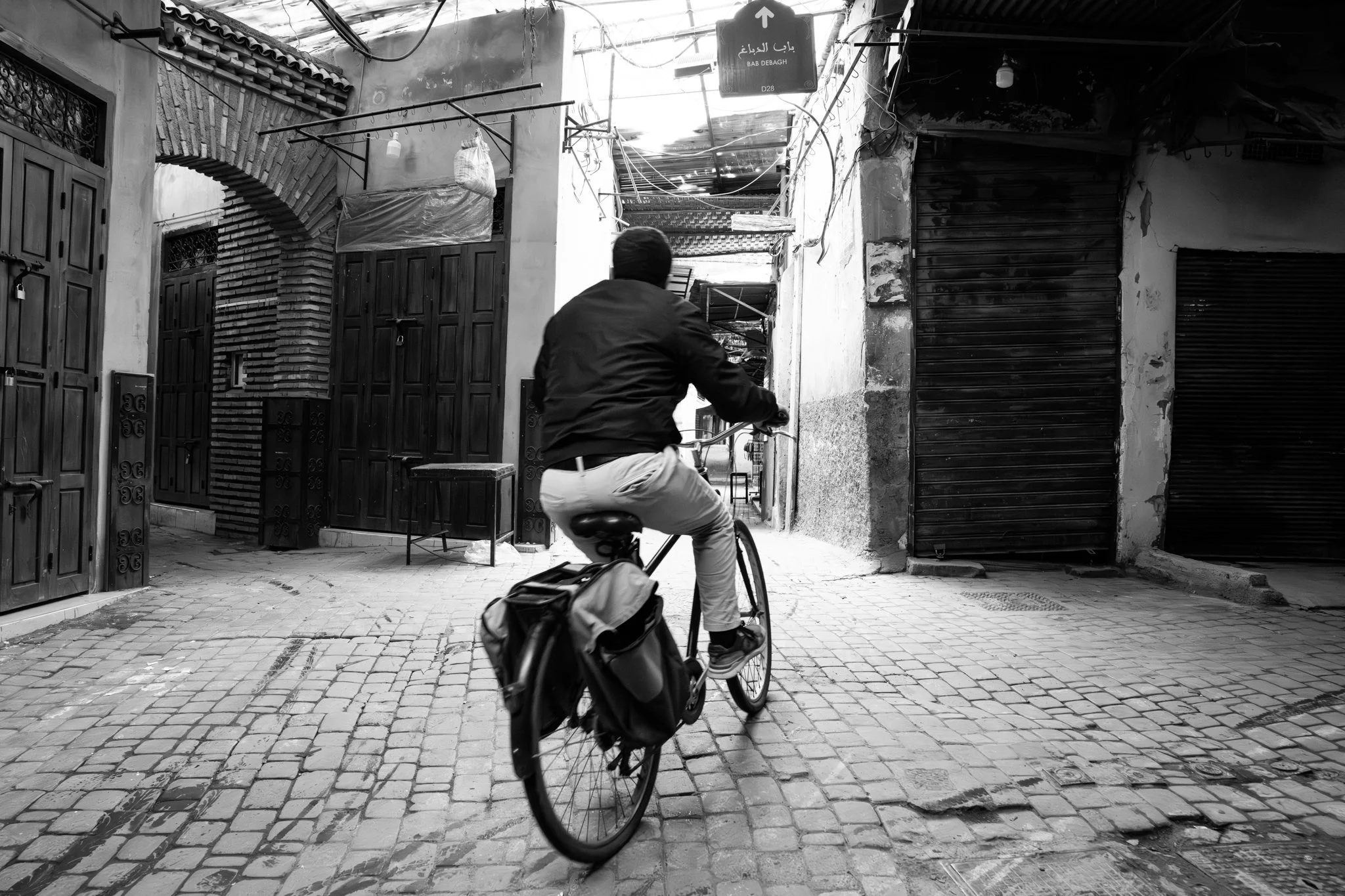 Echo Beneath the Stone, black-and-white fine art photograph by Jason Sponseller, featuring a cyclist moving through a Moroccan medina passage with architectural texture, shadow, and human presence