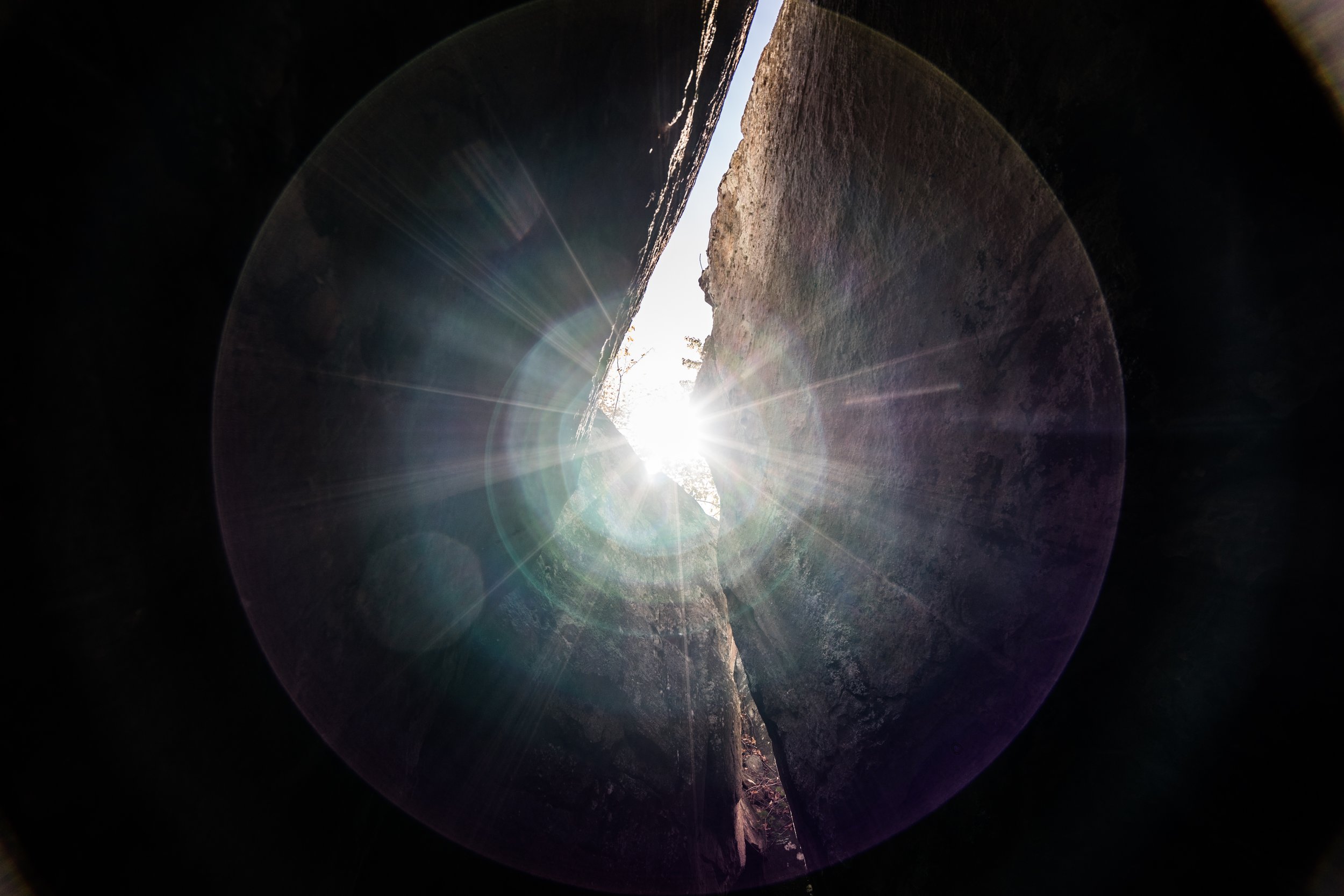 Dust and Distance: The Resonance of Time, limited edition fine art photograph by Jason Sponseller, featuring sunlight breaking through a narrow rock crevice with deep shadow, stone texture, lens flare, and a quiet sense of mystery