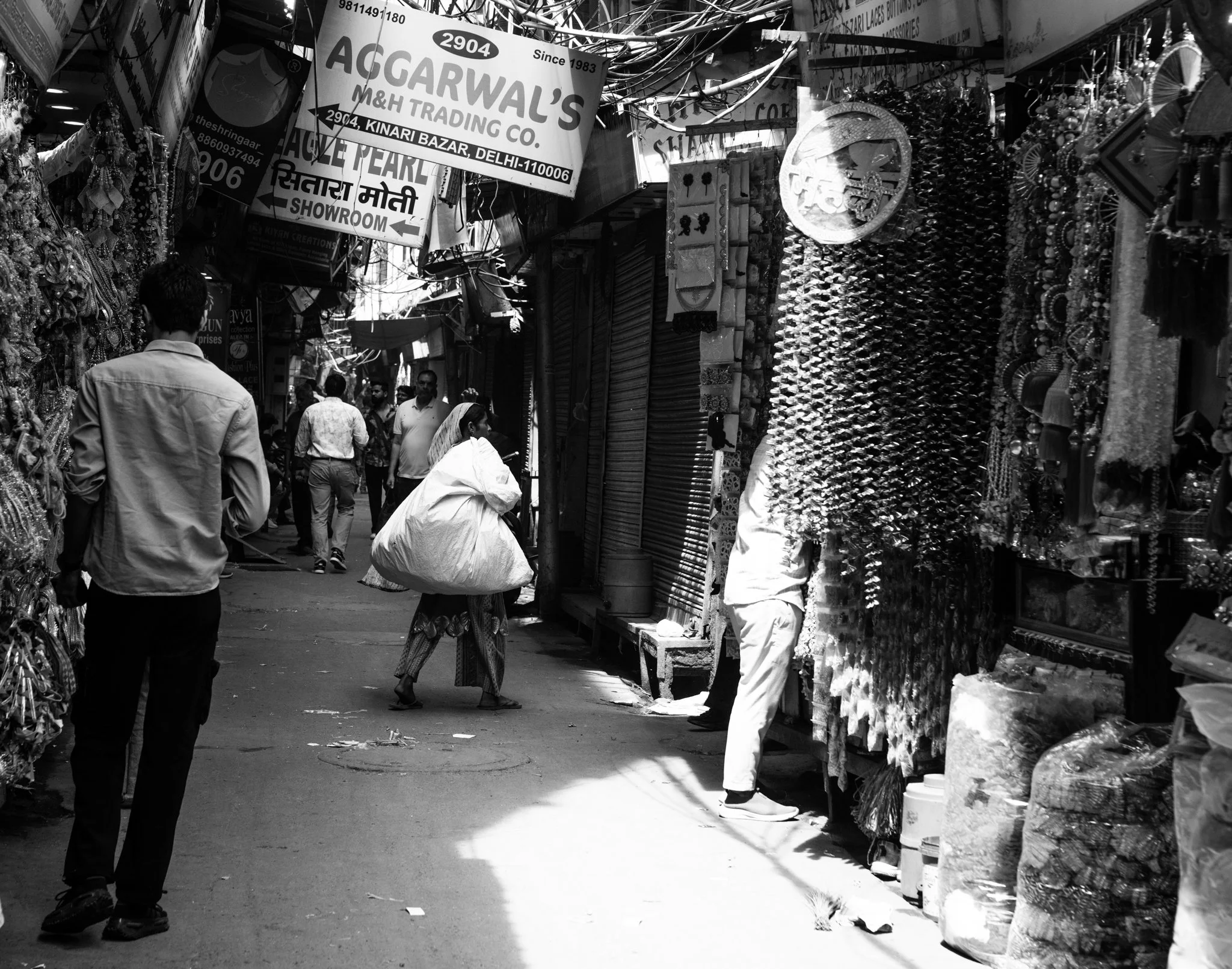 Echo Beneath the Stone, black-and-white documentary fine art photograph by Jason Sponseller, featuring Delhi’s Kinari Bazaar with layered market stalls, signage, shadow, and human presence