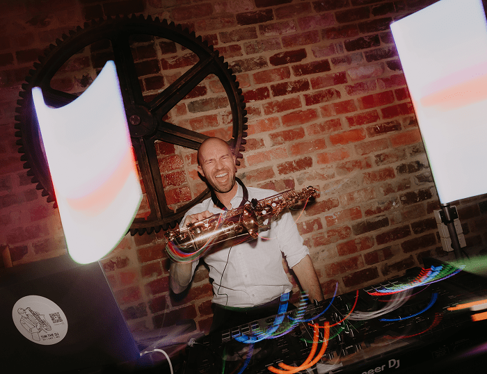 Tim the DJ performing with a saxophone, vibrant lighting, and DJ setup against a rustic brick wall.