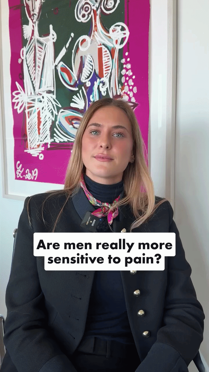 A woman sitting in front of a colorful abstract painting, wearing a black outfit and a pink patterned scarf around her neck, with the text 'Are men really more sensitive to pain?' overlayed on the image.