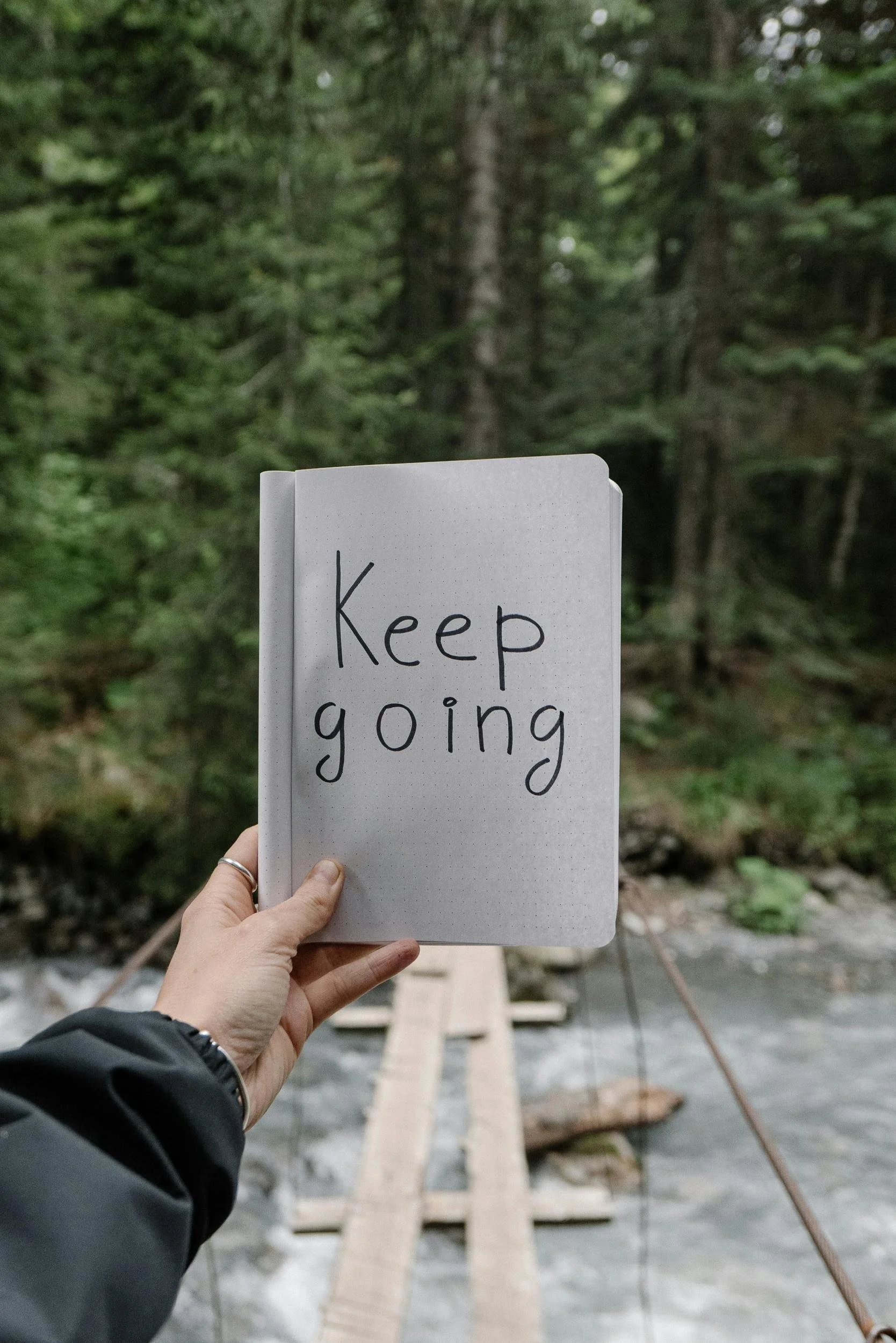 A stock photo of a hand holding a notebook with the words "Keep Going" in front of a wooded landscape.