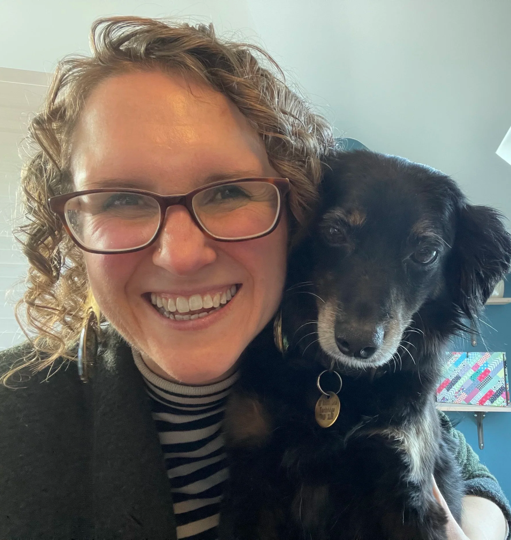 A woman with curly hair and glasses smiling with a black dog resting its head on her shoulder. The woman is wearing a striped shirt and a black sweater. The background includes some in-focus objects and a patterned colorful item.