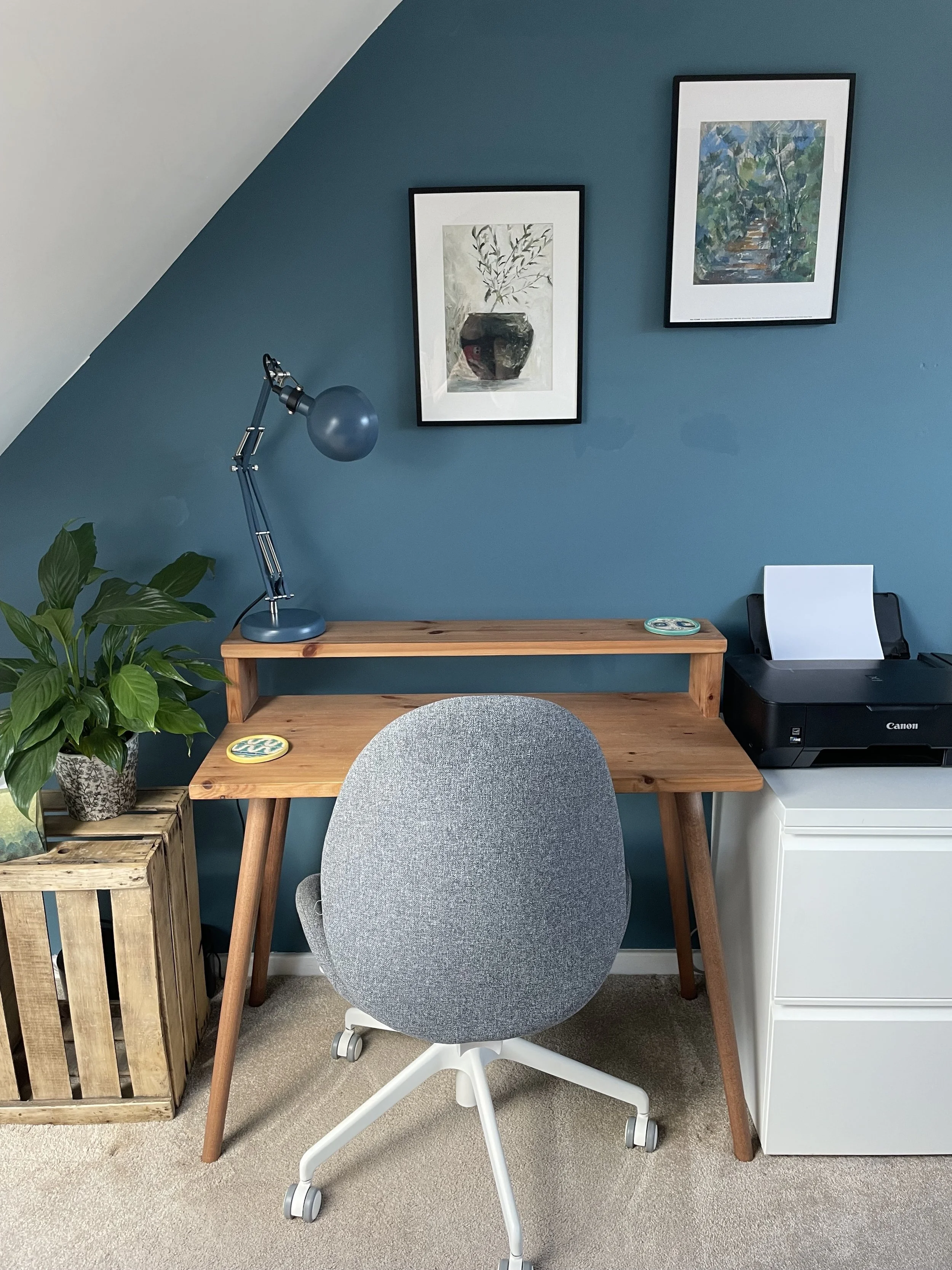 Home office with a wooden desk, gray swivel chair, green potted plant, blue wall with two framed paintings, desk lamp, and a printer.