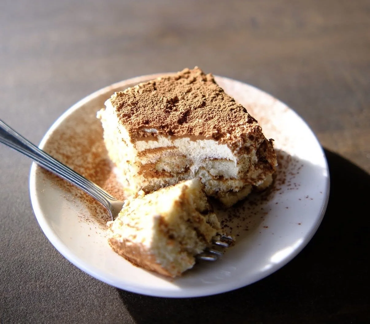 Close-up of a slice of tiramisu dessert in a white bowl, with a spoon partially inside, topped with cocoa powder.
