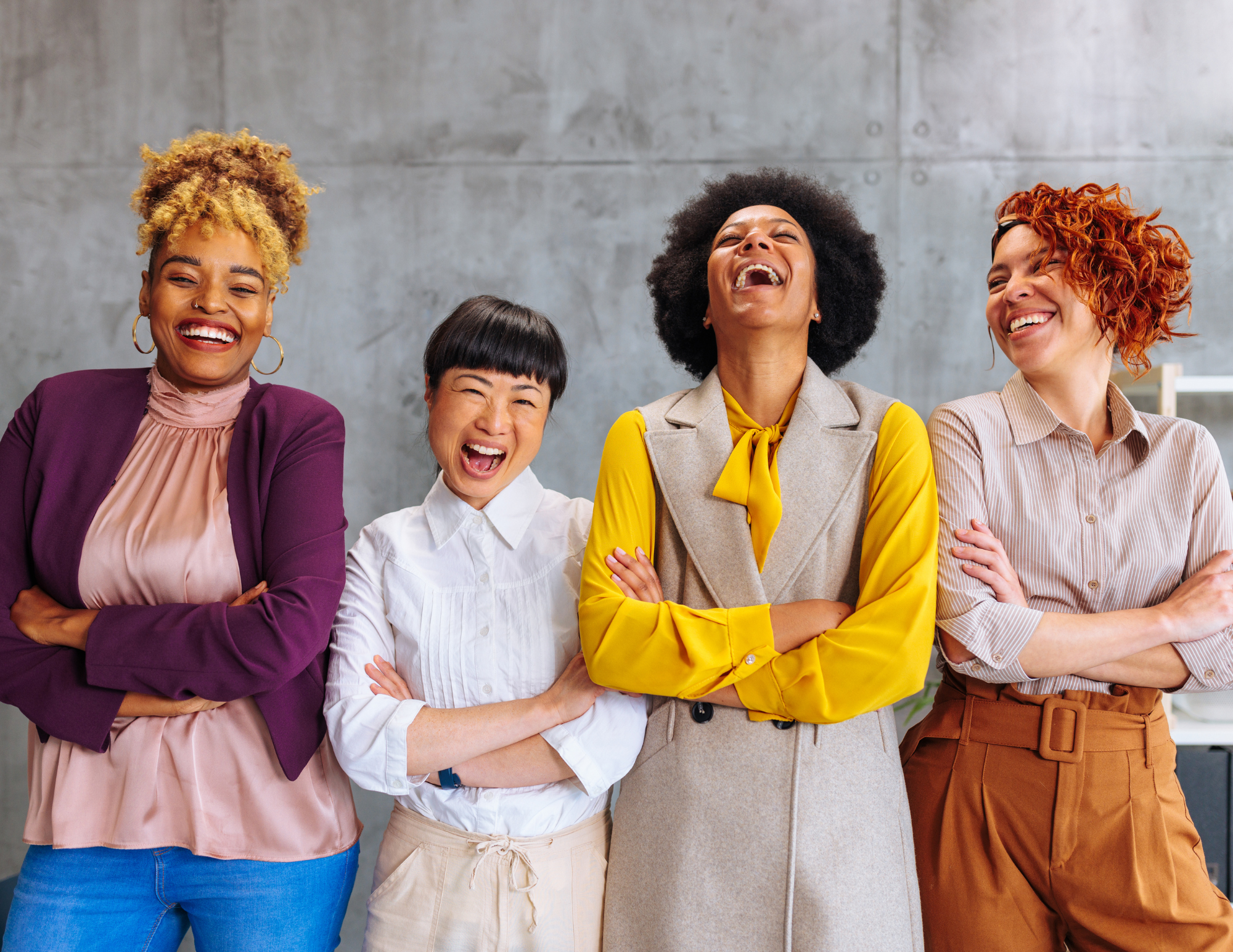 Four women standing together in an office, laughing and smiling, wearing colorful professional outfits.