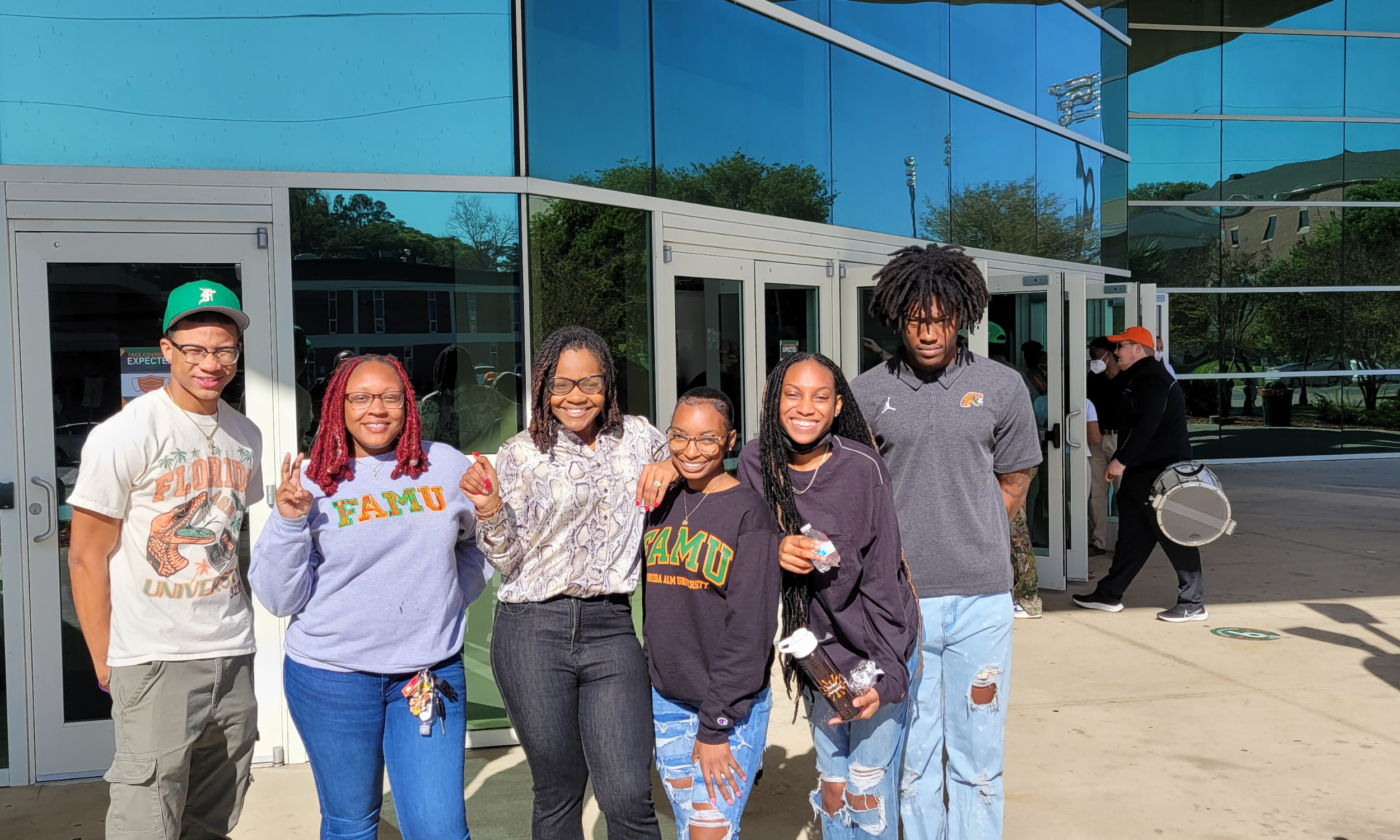Group of six young adults standing outside in front of a glass building, some wearing Florida A&M University apparel, smiling and posing for the camera.
