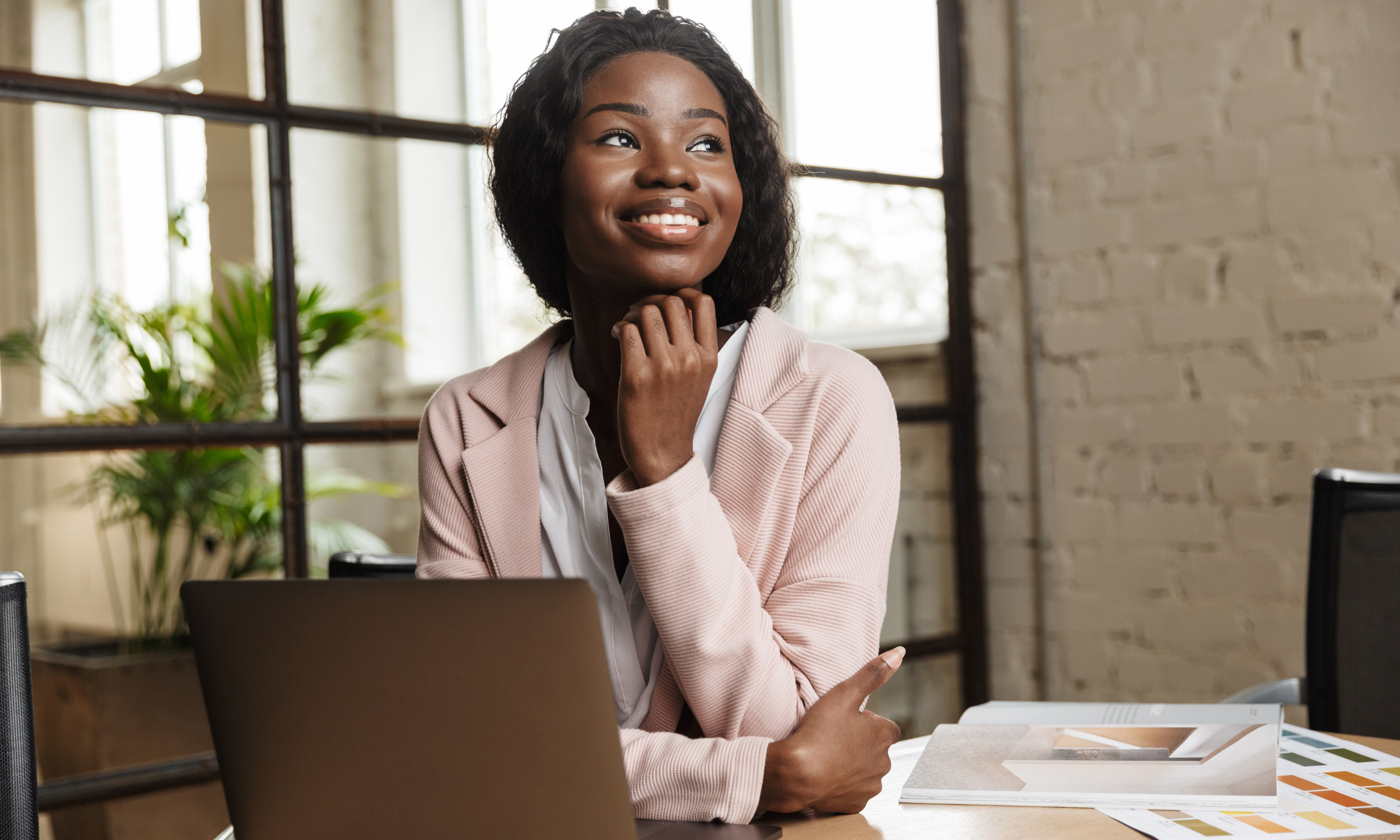 A woman with dark skin and curly hair smiling and resting her chin on her hand, sitting at a desk with an open laptop and design samples in a bright office.