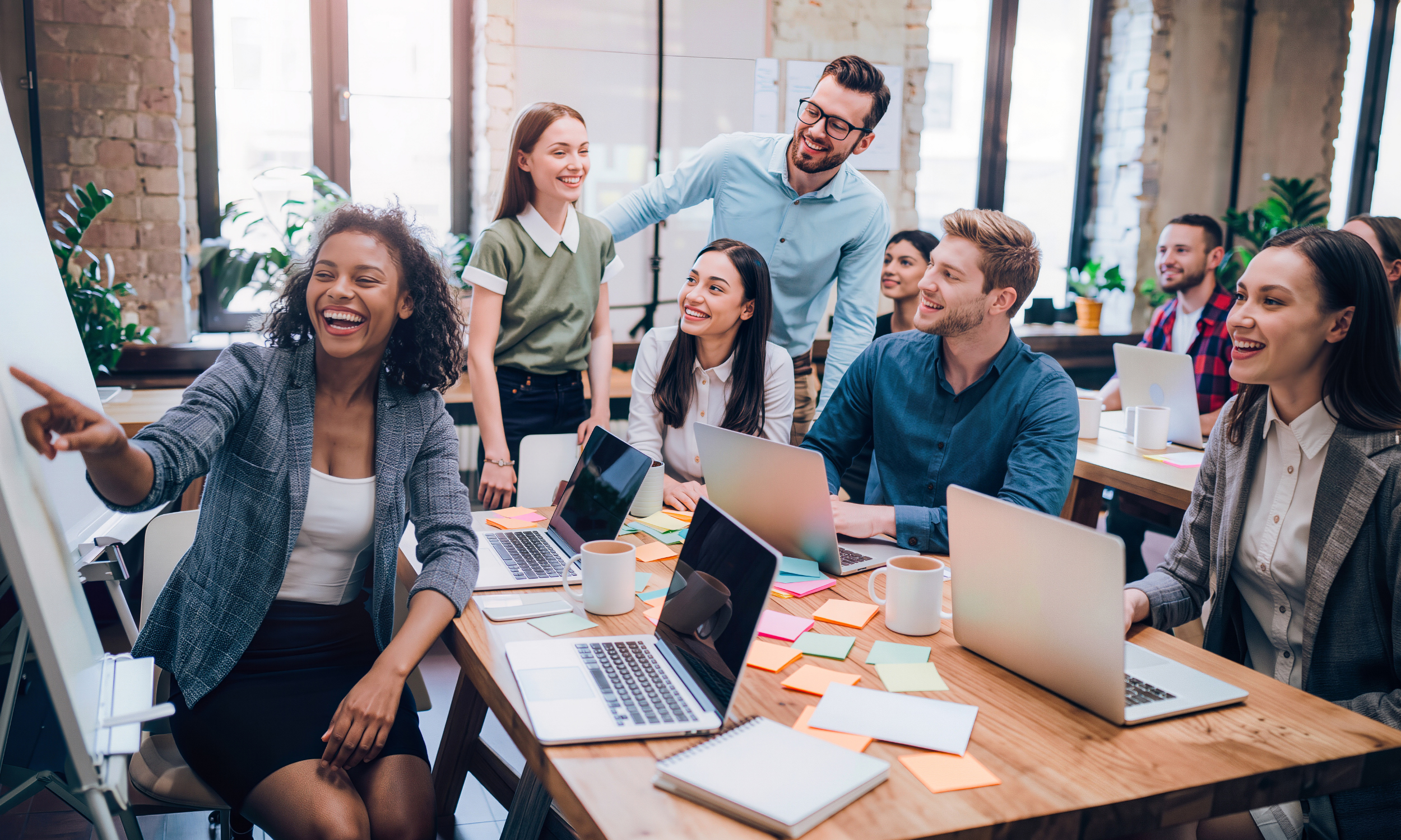 Group of colleagues in a modern office during a meeting, with one woman pointing at a whiteboard and others smiling, surrounded by laptops, notebooks, and coffee mugs.