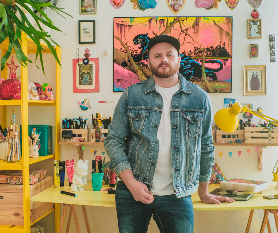A man with a beard wearing a denim jacket and a black cap standing in a colorful, creatively decorated room with artwork, crafts, and art supplies.