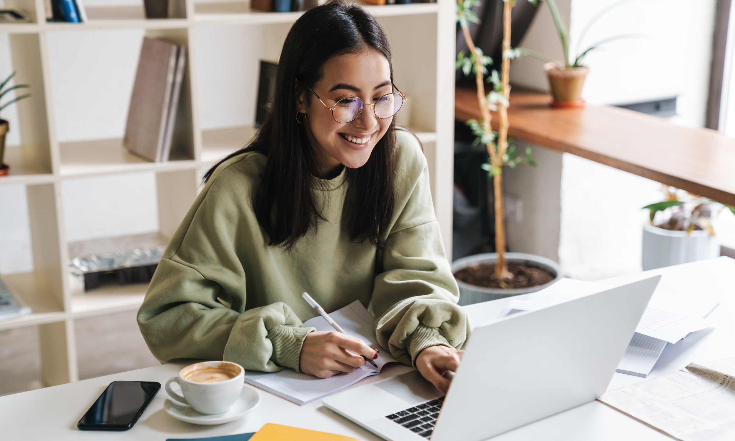 A young woman with glasses and dark hair smiling while working on a laptop, surrounded by notebooks, a smartphone, a cup of coffee, and papers in a bright, modern office or cafe setting.