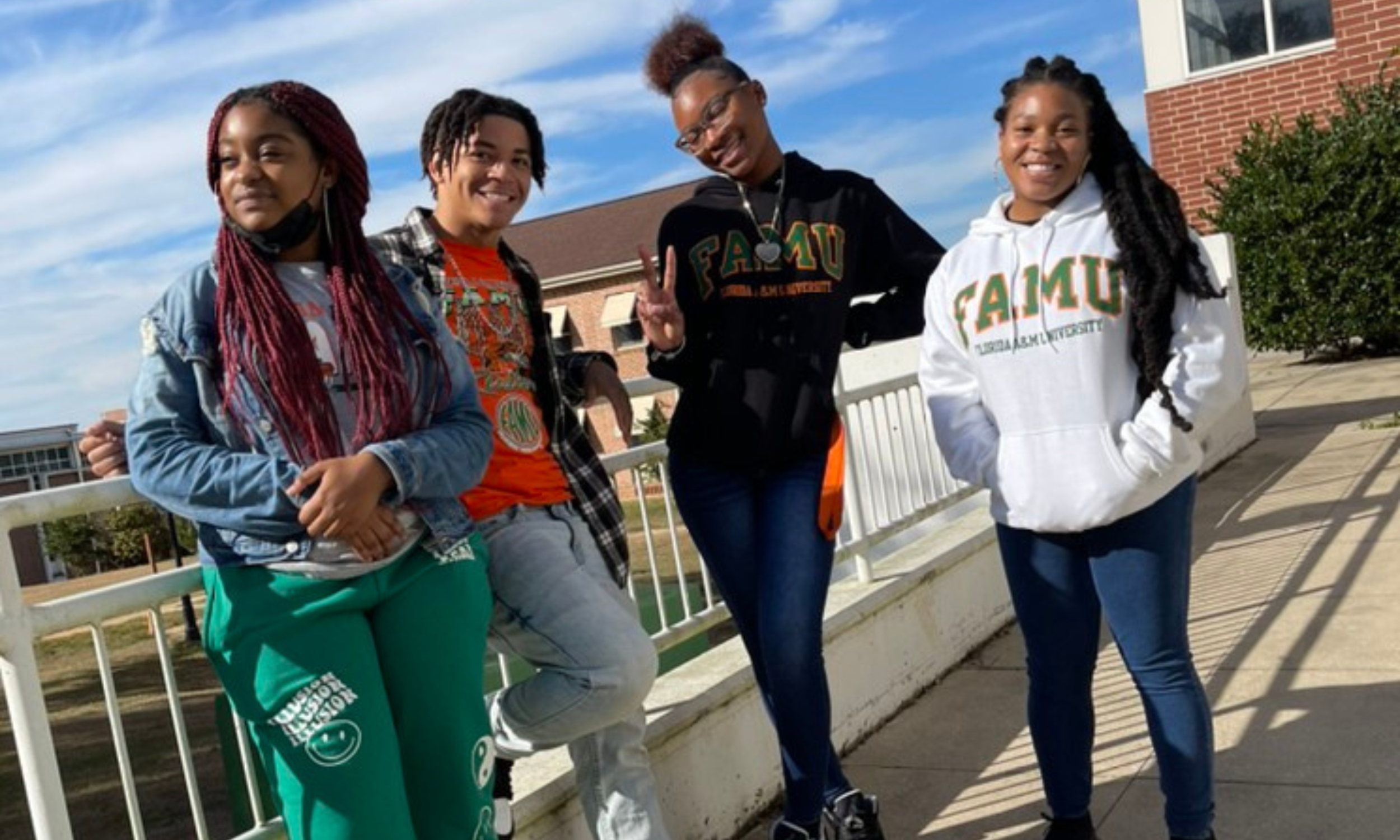 Four diverse young women standing outside on a sunny day, smiling, with a brick building and greenery in the background. Three women are wearing university sweatshirts that say 'FAMU'.