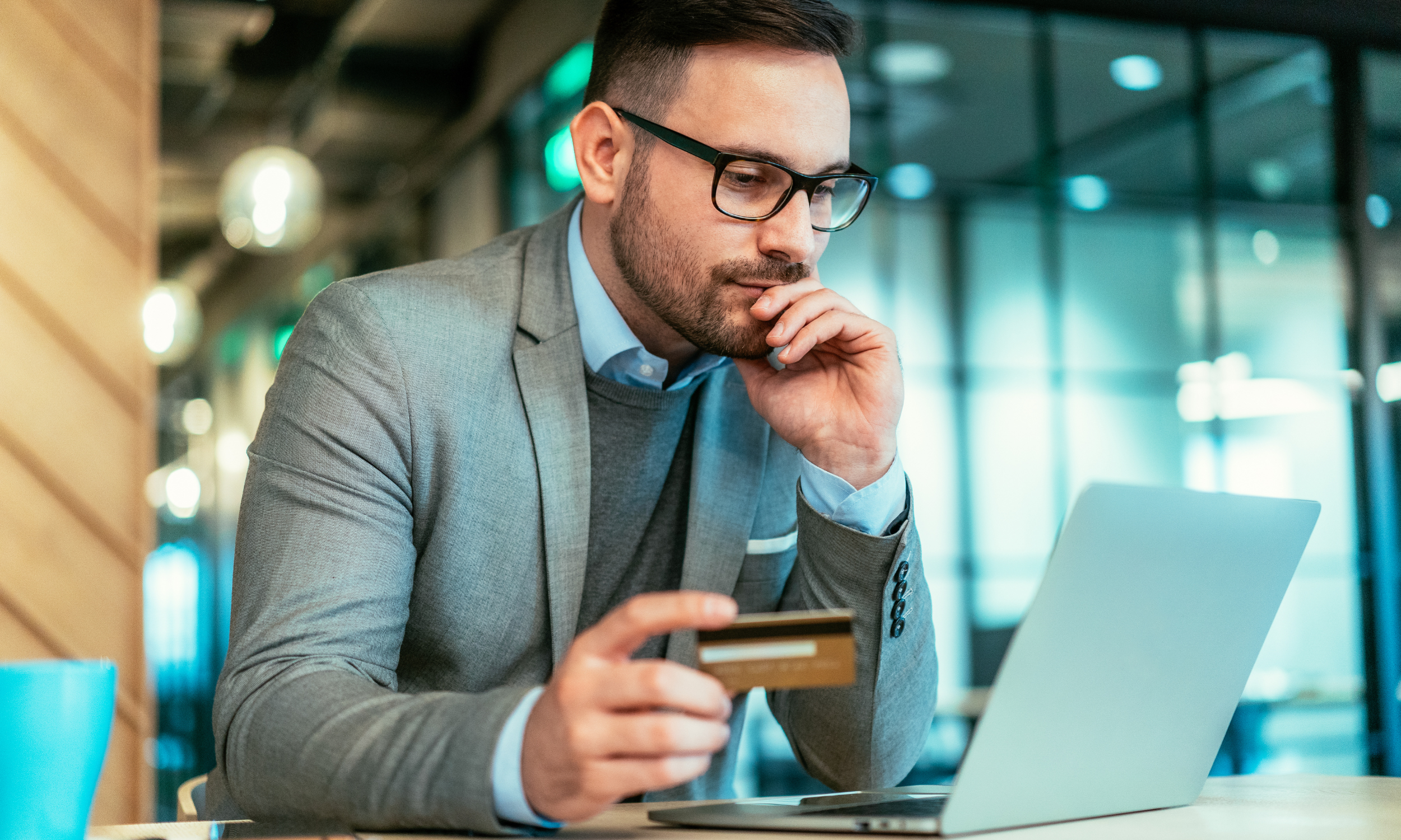 Man in glasses and gray suit holding a credit card and looking at a laptop screen in a modern indoor space.