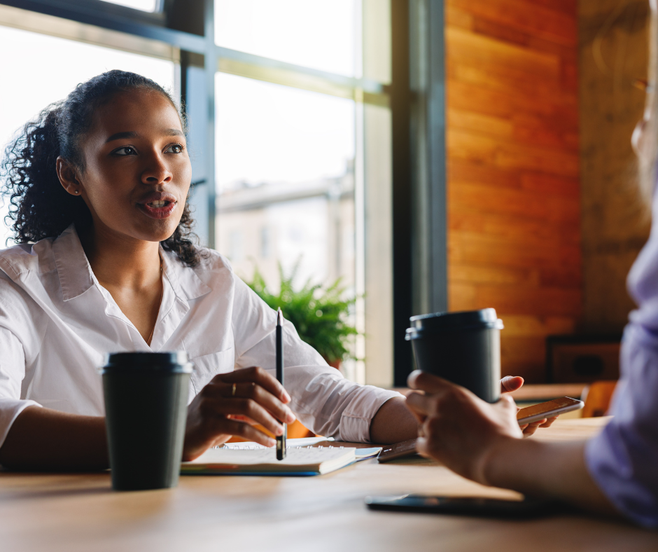 Two women having a discussion in a coffee shop, with coffee cups, notebooks, and a smartphone on the table.