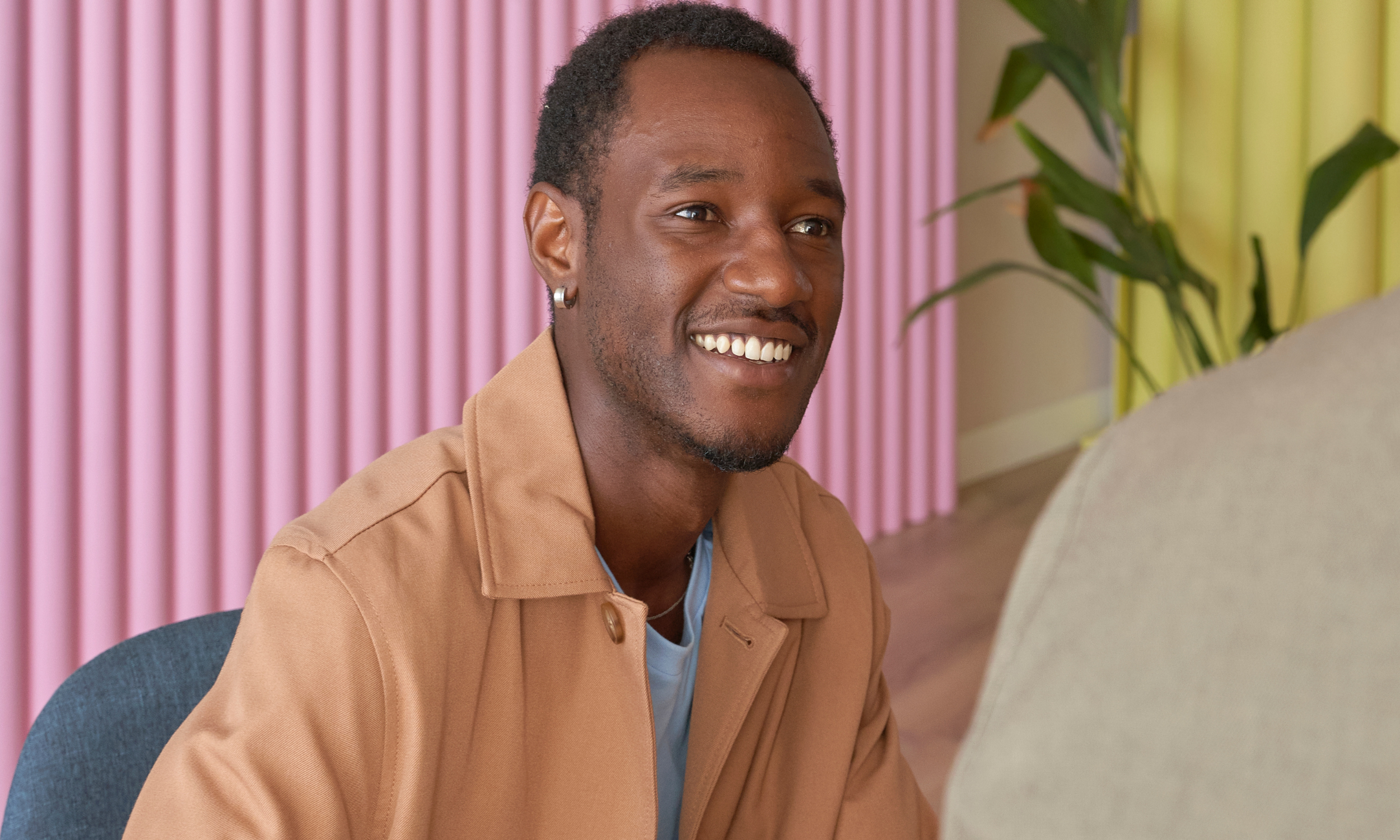 A smiling man with dark skin, short curly hair, piercings, and wearing a tan jacket, seated indoors in front of pink and yellow walls with a green plant nearby.