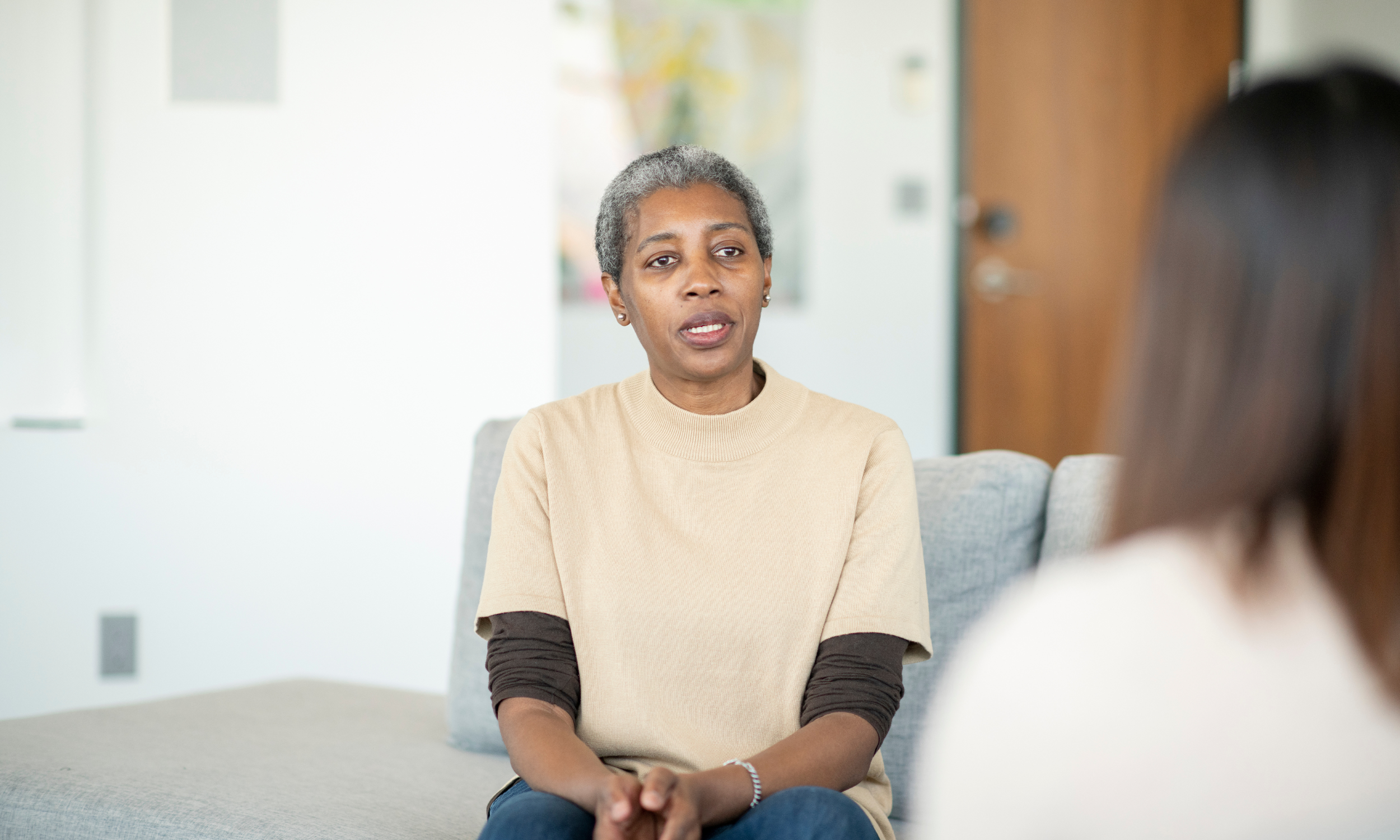 A woman with short gray hair and dark skin sitting on a gray couch, talking to a person with long dark hair, indoors with a white wall and doorway in the background.