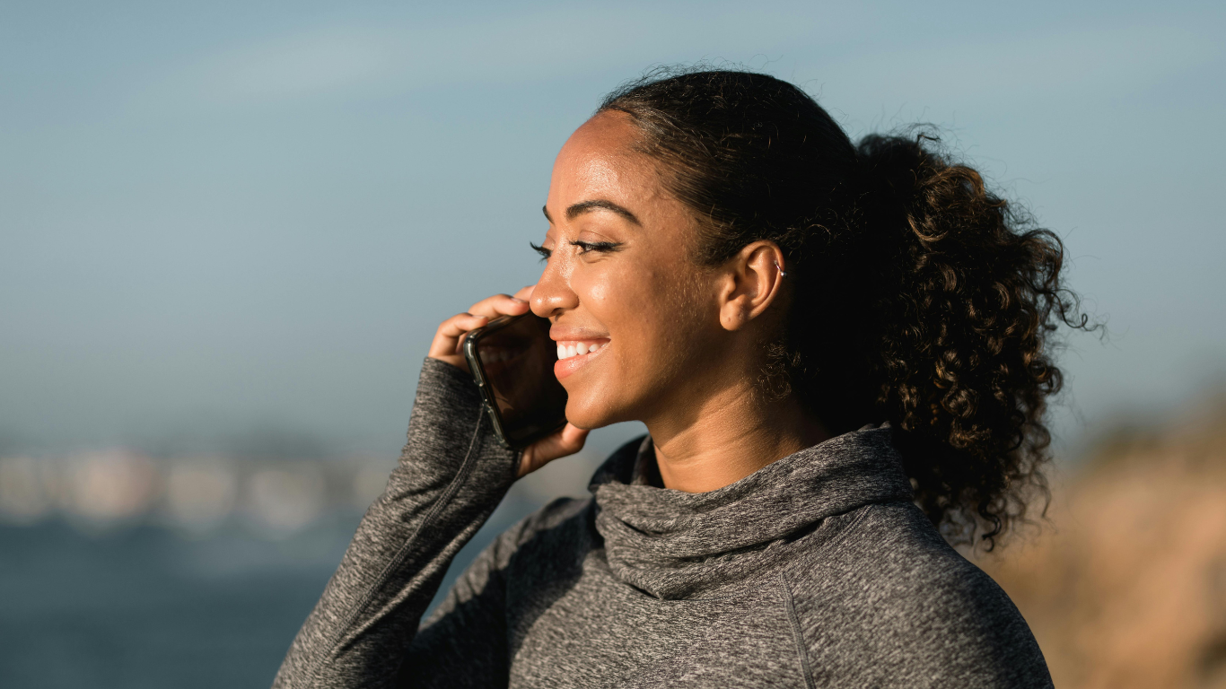 A woman with curly hair smiling while talking on her cellphone outside, wearing a gray athletic top.