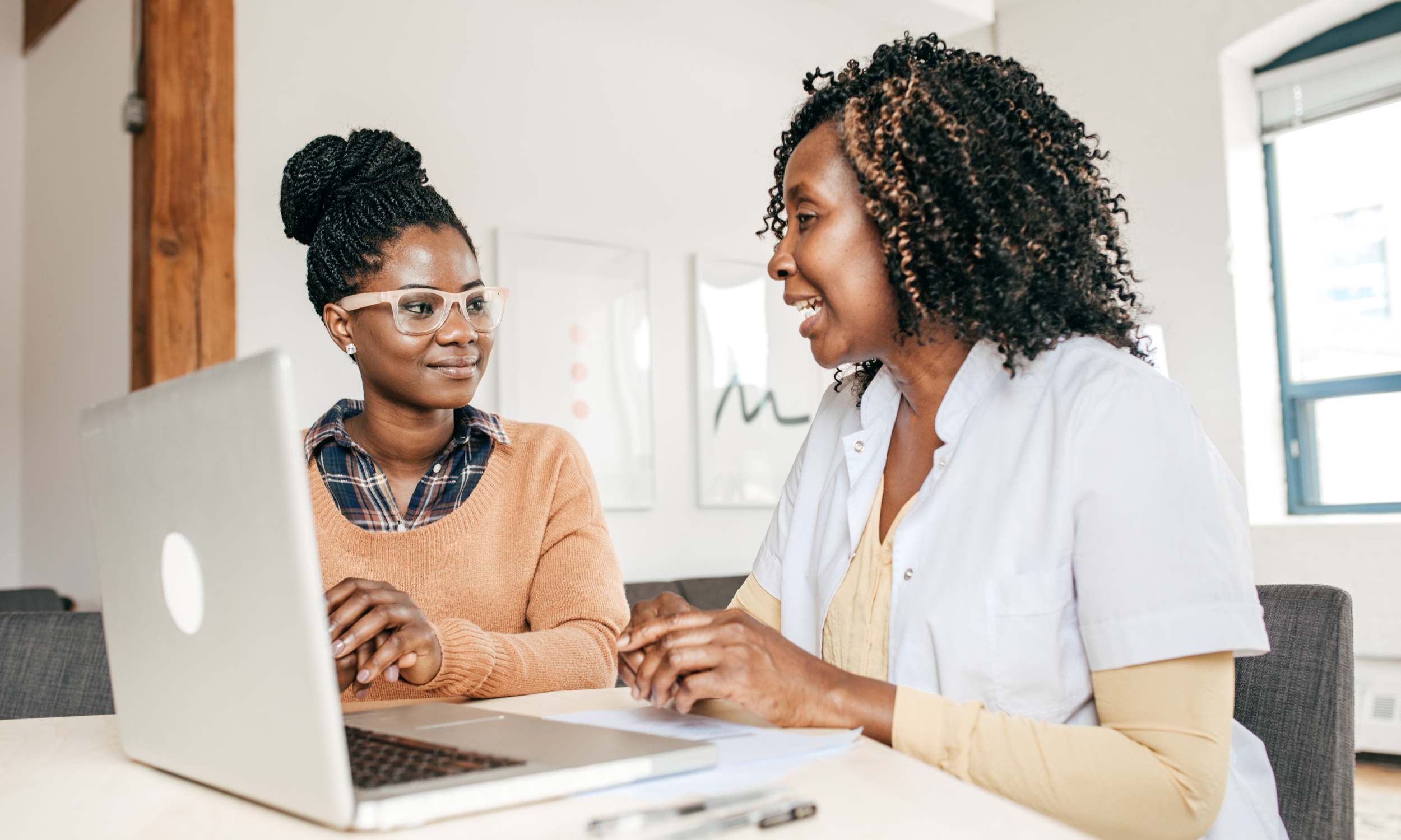 Two women sitting at a table in an office, having a conversation. One woman is wearing glasses and a plaid shirt under a tan sweater, while the other woman is wearing a white shirt and smiling. A silver laptop is on the table.