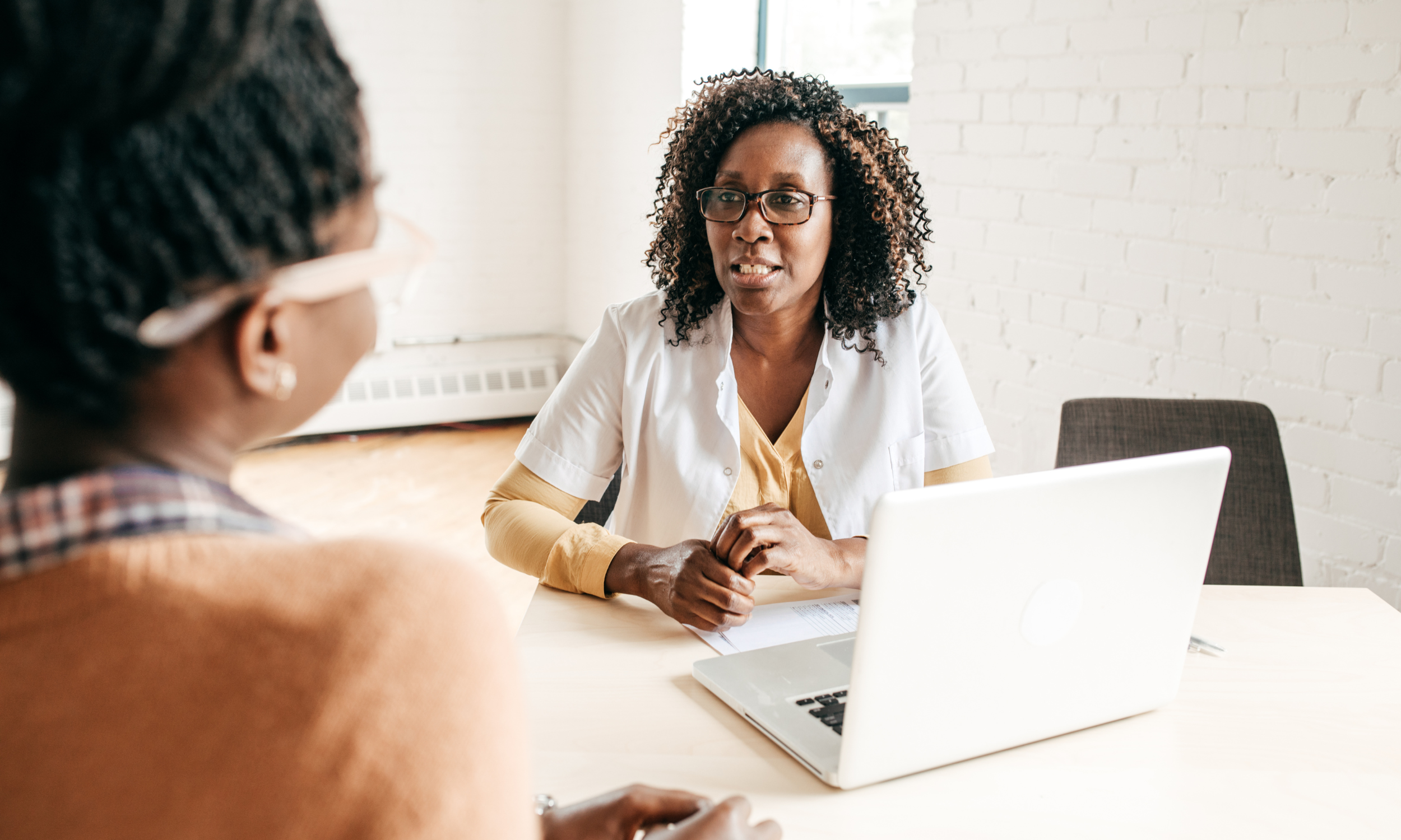 Two women having a conversation in an office, one woman is talking and the other listening, with a laptop on the table between them.