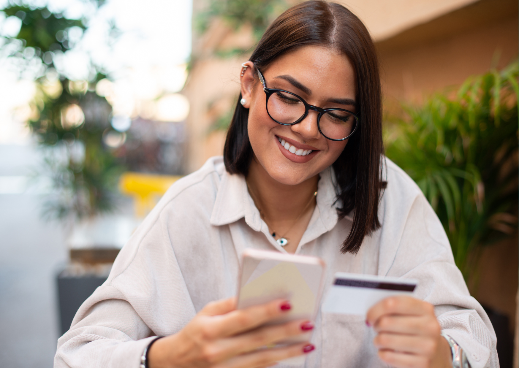 A smiling woman with glasses holding a smartphone and a credit card, sitting outdoors with plants in the background.