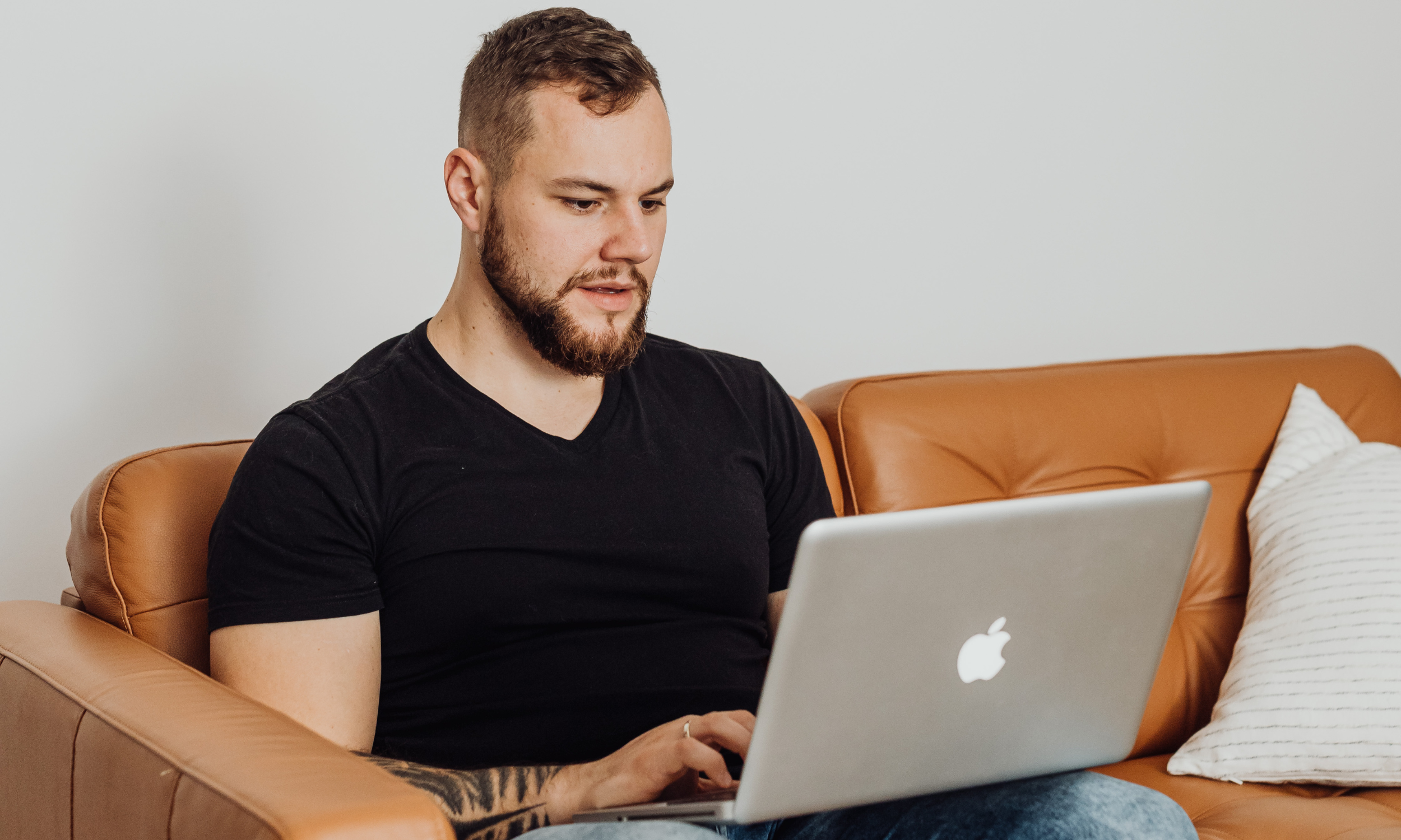A man with a beard and wearing a black t-shirt sitting on a tan leather sofa, working on a silver MacBook laptop.