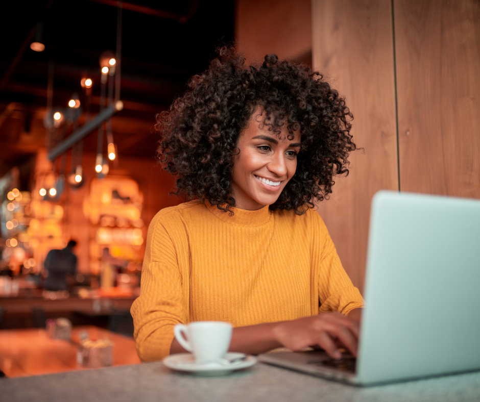 A woman with curly hair wearing a yellow sweater sitting at a table with a laptop and a white coffee cup, smiling in a warmly lit cafe.
