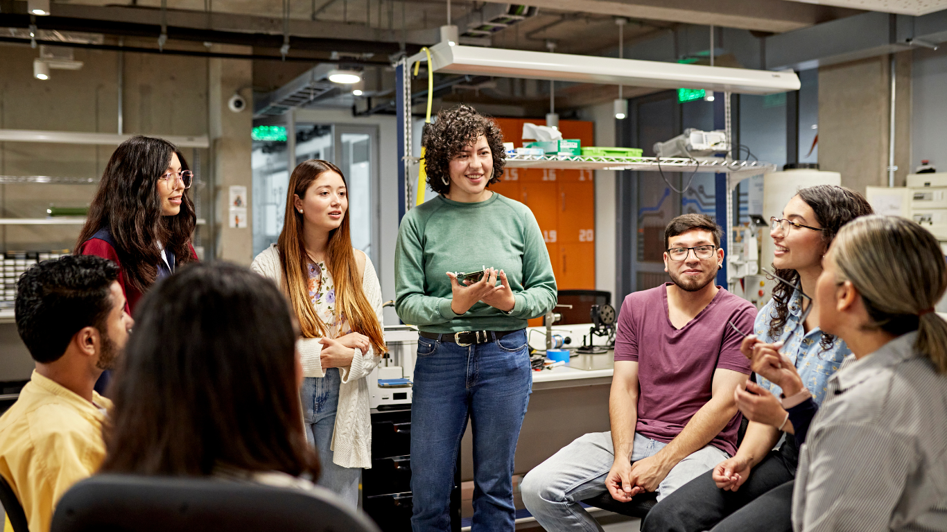 A diverse group of young adults having a meeting or discussion in a modern office or laboratory space, with some standing and others sitting, engaging in conversation and listening.