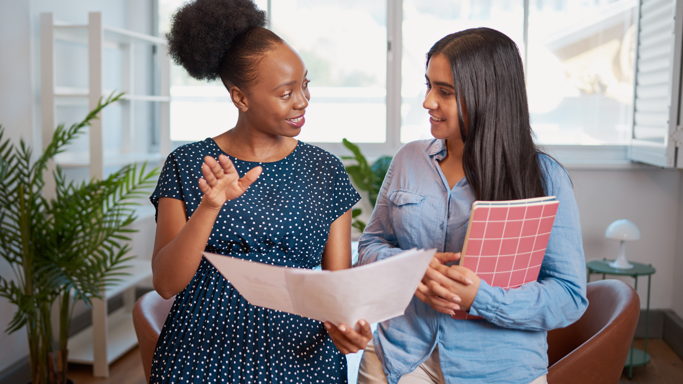 Two women are having a friendly conversation indoors. One woman, with dark skin and wearing a navy polka dot dress, is holding a sheet of paper and gesturing with her hand. The other woman, with light skin and long dark hair, is holding a pink and white checkered notebook and smiling at her. There are green plants and a window in the background.