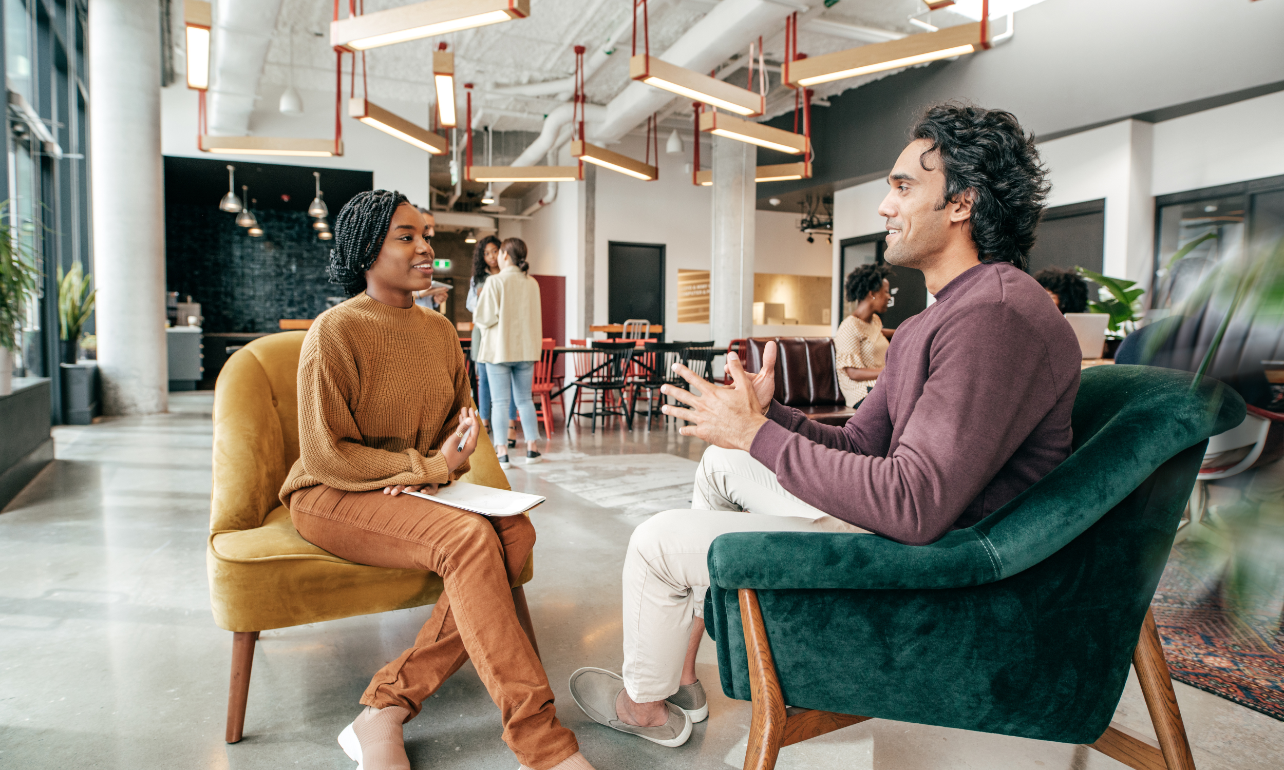 A man and woman having a conversation in a modern, well-lit cafe with other people in the background.