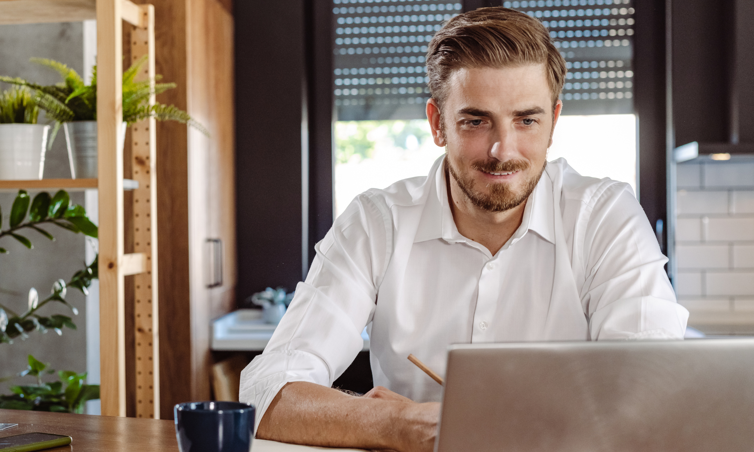 A man with light brown hair and a beard smiling while working on a laptop at a wooden table in a modern room with plants and black window blinds.