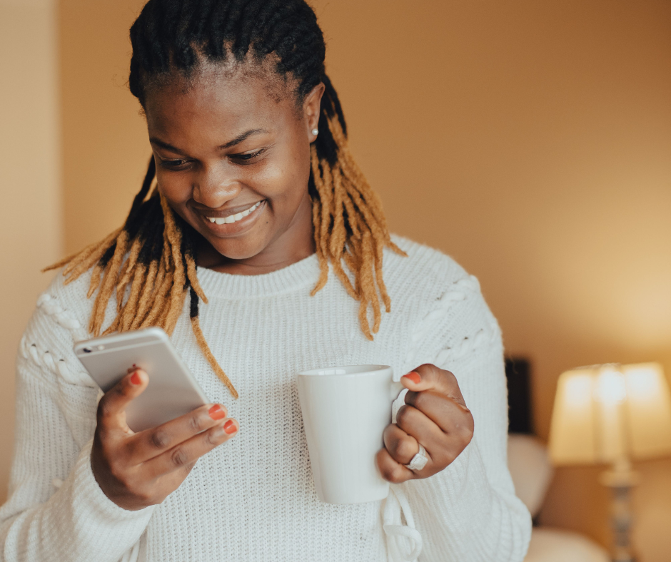 A woman with dreadlocks smiling while looking at her phone and holding a white mug indoors with warm lighting.