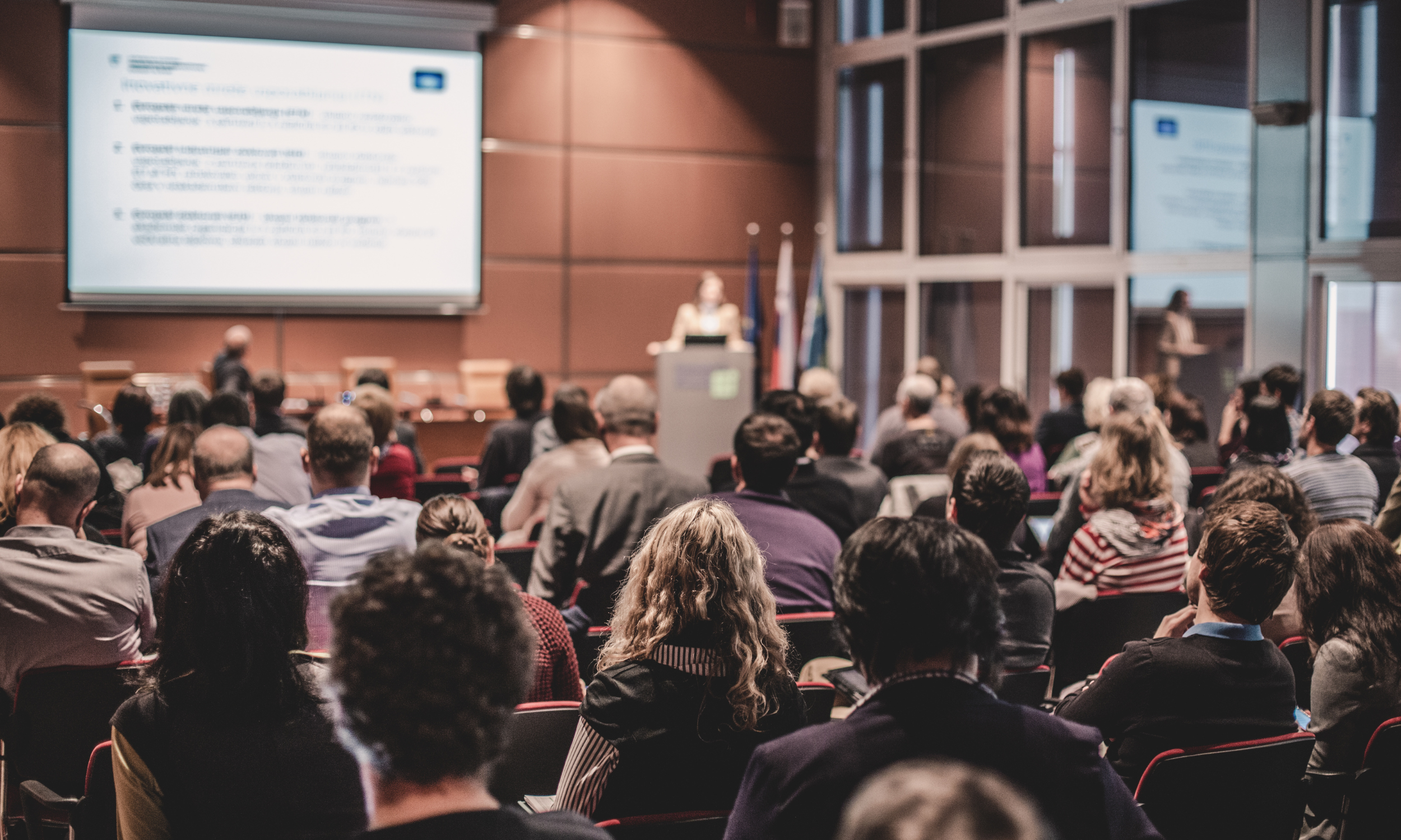 Indoor conference room filled with seated attendees, facing a speaker at a podium, with a large presentation screen displaying text in the background.