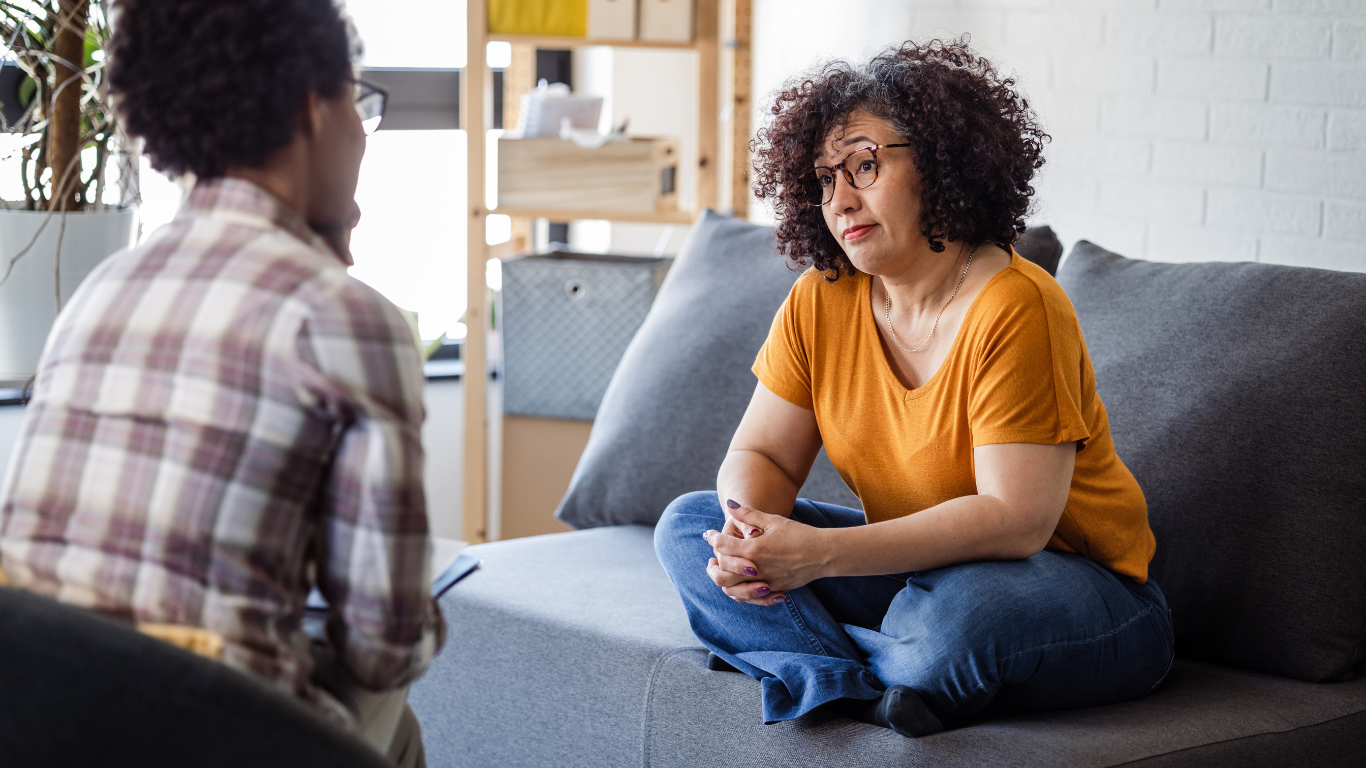 A woman with curly hair, glasses, and an orange shirt sitting cross-legged on a gray couch, listening attentively to another person who is taking notes.