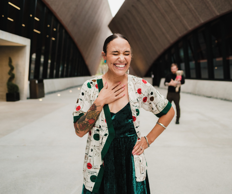 A young woman with dark hair smiling with her hand on her chest, wearing a green dress and a patterned jacket, standing outdoors in an urban setting with a modern building in the background, and another woman holding a dog in the distance.