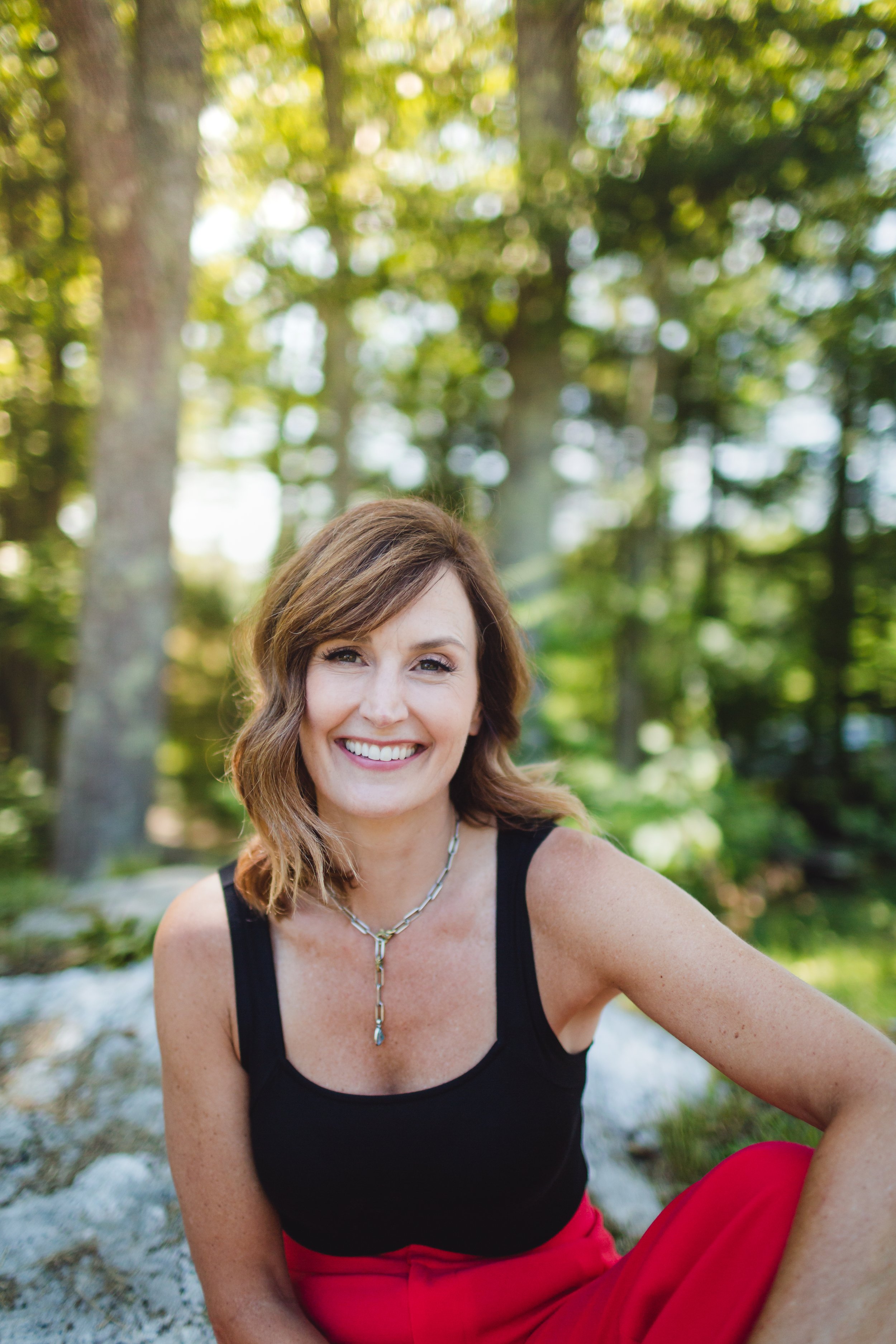 A woman with shoulder-length brown hair smiling outdoors in a green forest, wearing a black tank top, red pants, and a silver necklace.