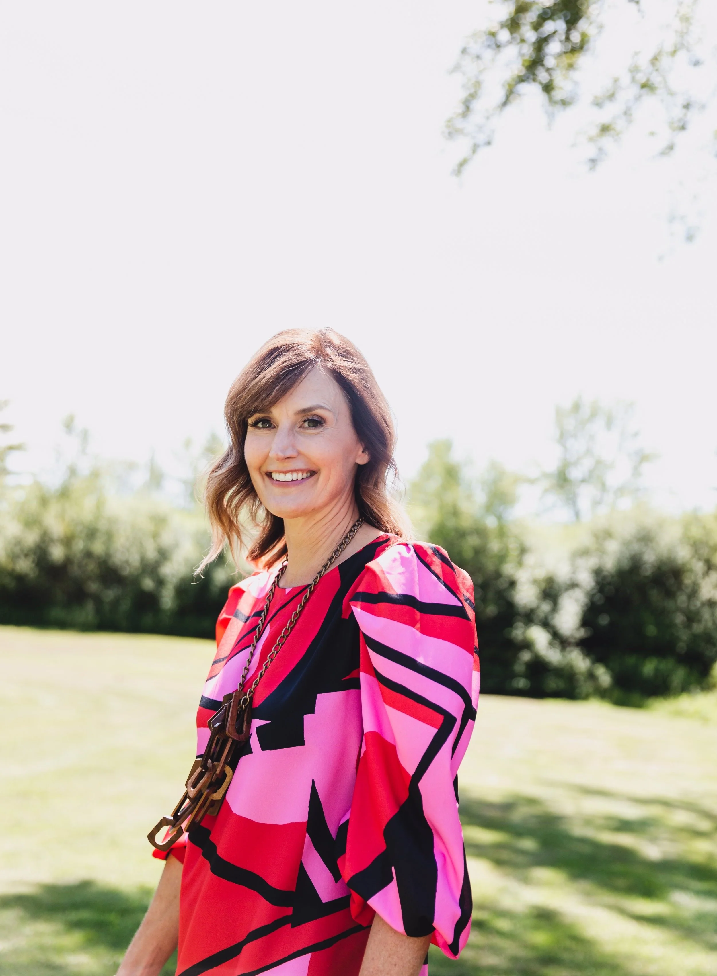 A woman wearing a colorful pink and red patterned dress standing outdoors in a grassy area with trees in the background, smiling at the camera.