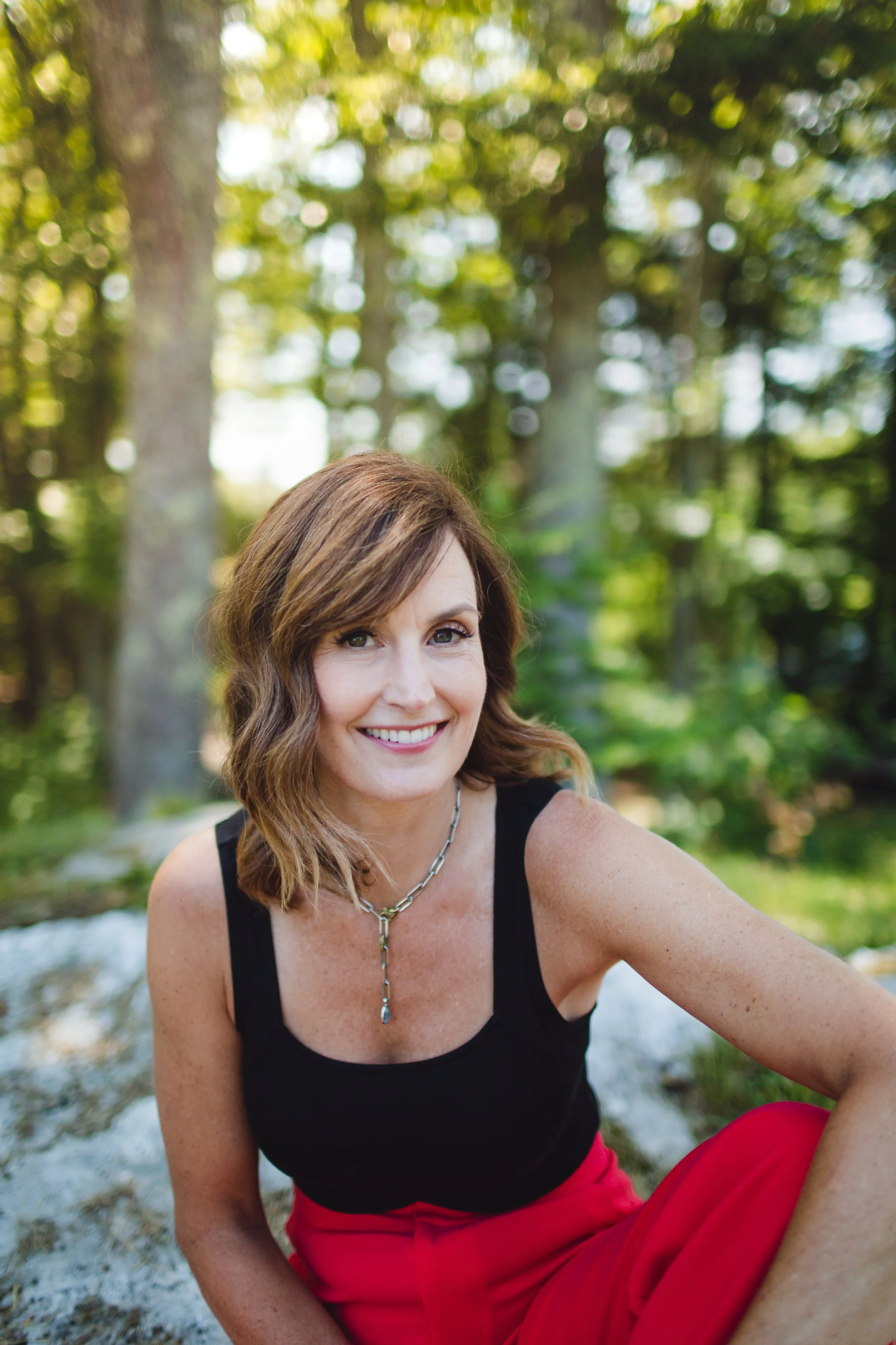 A woman with shoulder-length brown hair wearing a black sleeveless top and red pants sitting outdoors in a forested area, smiling at the camera.