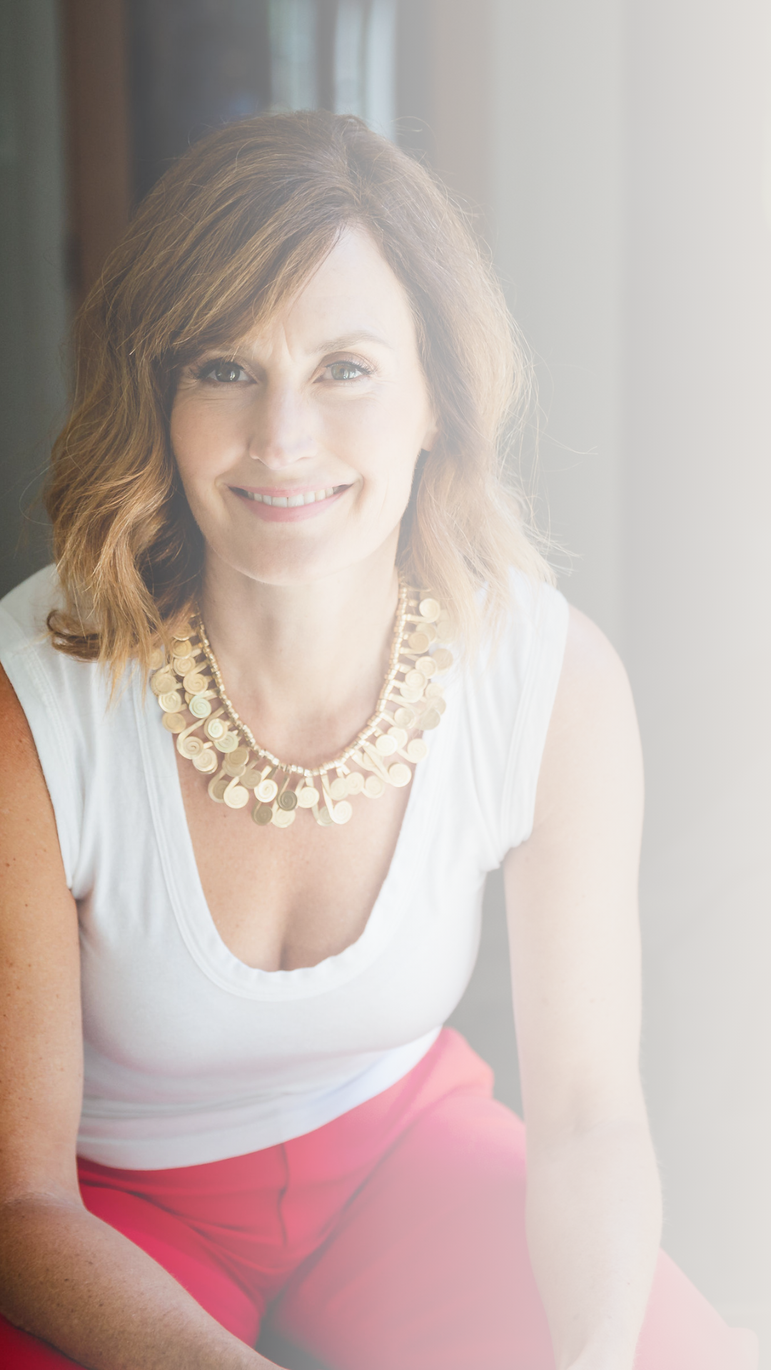 A smiling woman with shoulder-length brown hair, wearing a white sleeveless top, a gold necklace, and pink pants, sitting by a window with sunlight.