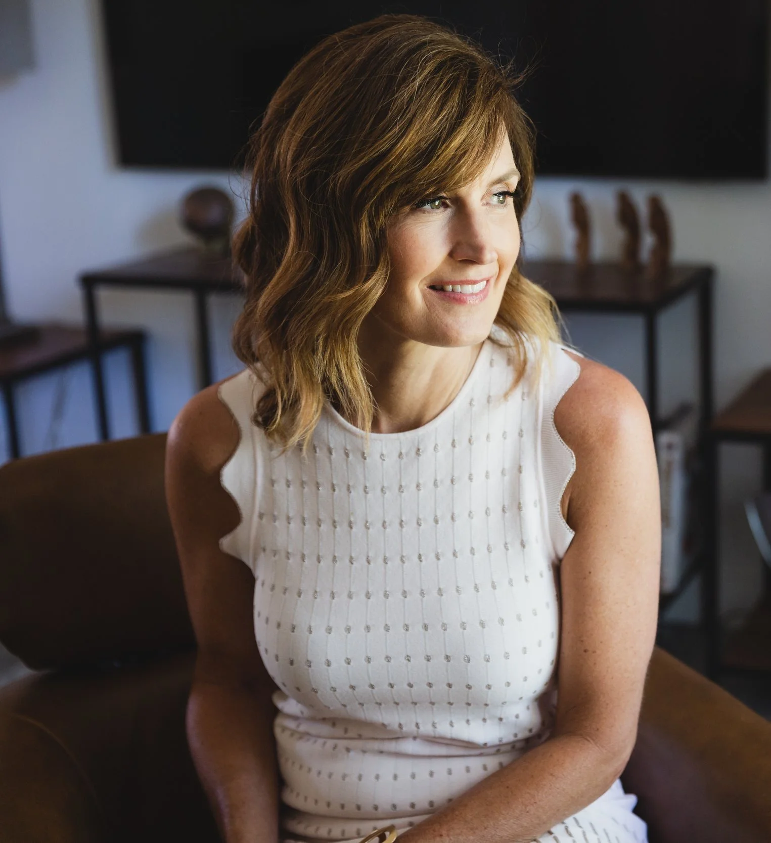 A woman with wavy, shoulder-length hair sitting on a brown chair, smiling and looking to her right, in a room with dark furniture in the background.