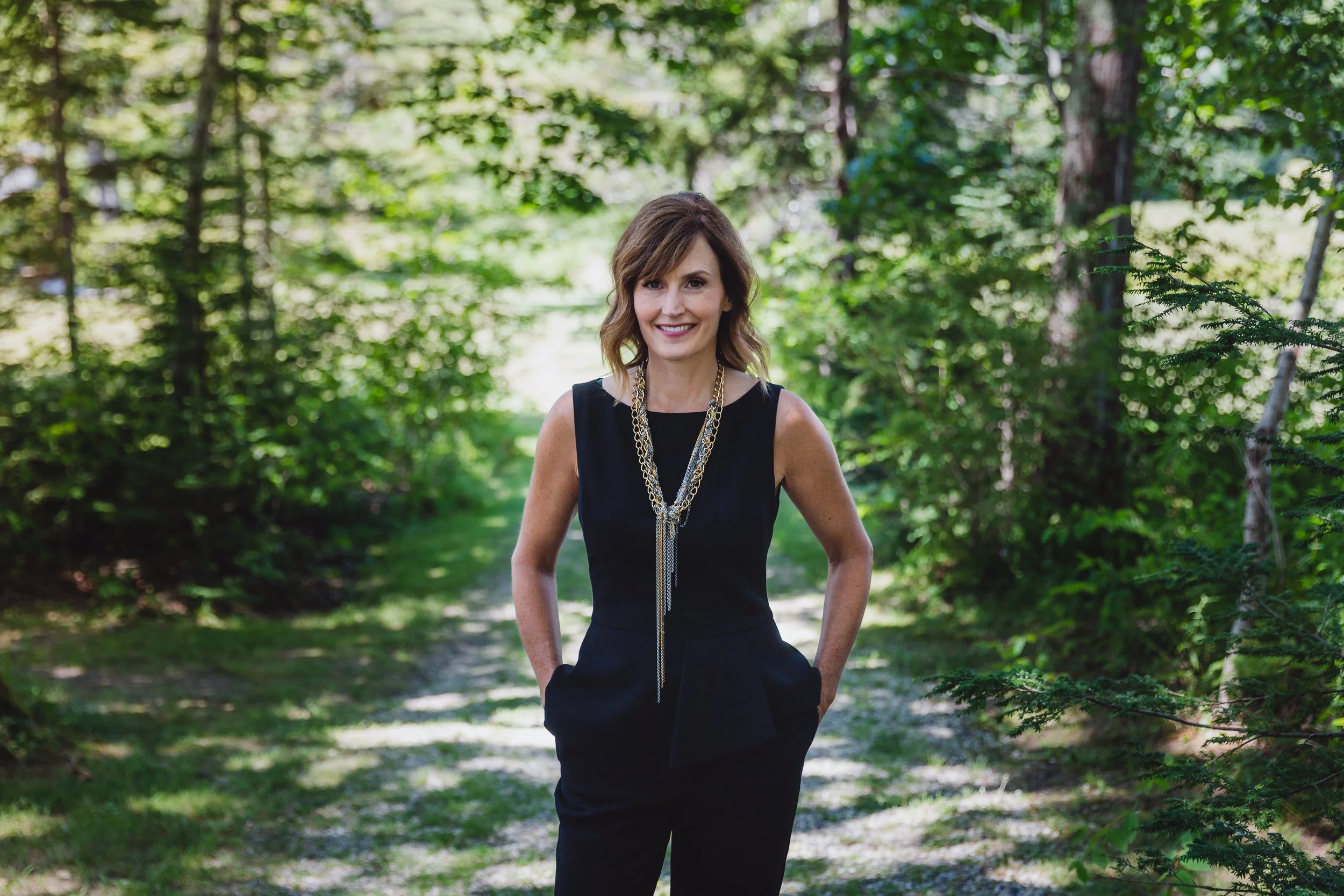 A woman standing on a forest path, wearing a black sleeveless top and dark pants, with a long gold necklace and smiling at the camera.