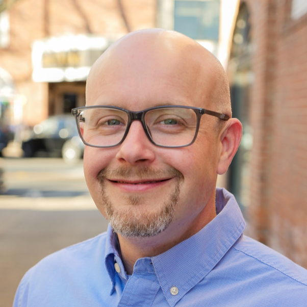 A smiling man with glasses, a goatee, and a shaved head, wearing a blue button-up shirt, standing outdoors with a brick building and cars in the background.