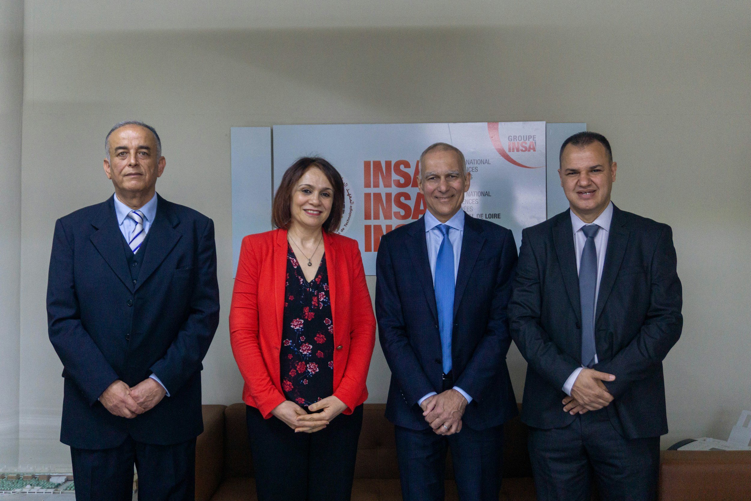Four professionally dressed individuals standing in front of a sign that reads "INSA" and related text. The group includes two men in suits, a woman in a red blazer, and a man in a dark suit and tie.