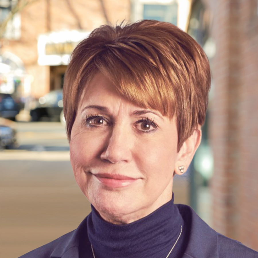 Close-up of a woman with short, reddish-brown hair and a friendly smile, wearing a navy blazer and a black turtleneck, outside in an urban setting.