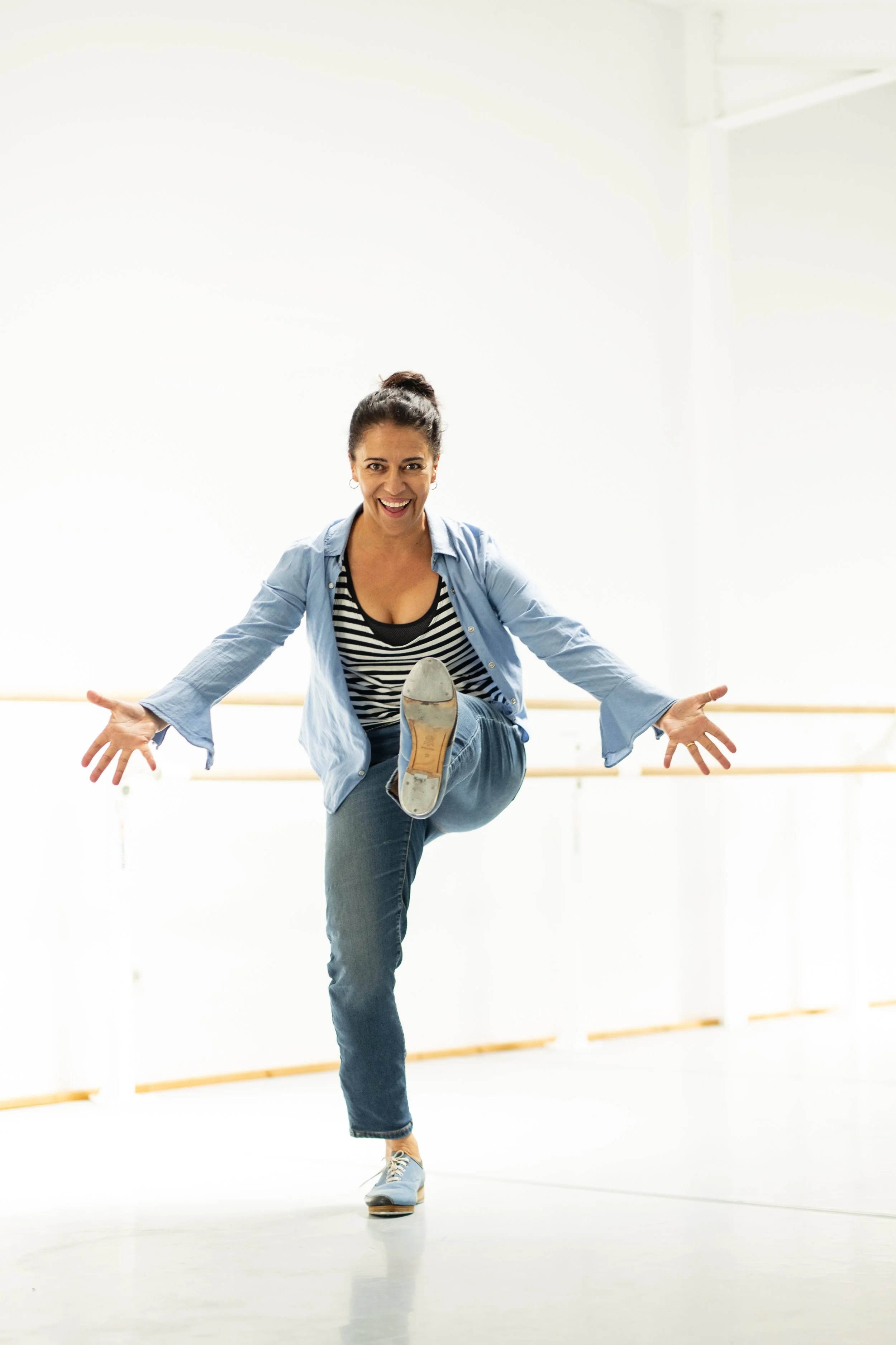 Une femme souriante en tenue décontractée, levée d'une jambe et bras ouverts, dans une salle de danse ou studio de ballet