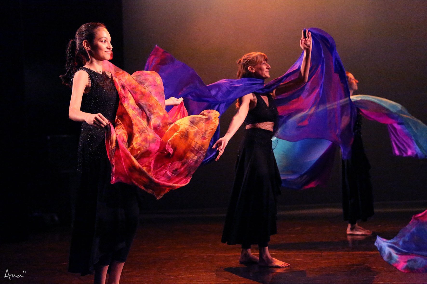 Des femmes danseuses en costumes noirs tiennent des tissus colorés lors d'une performance de danse sur scène. La scène est sombre avec un éclairage chaud.