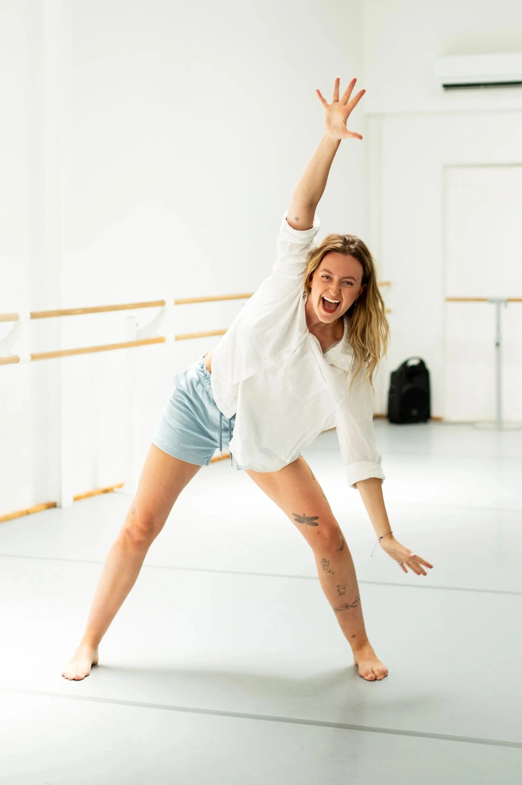 Femme souriante dans une salle de danse, en position de danse ou d'exercice, portant un t-shirt blanc, bermuda court, avec des tatouages sur les jambes, dans une pièce lumineuse avec des barres de danse sur le mur.