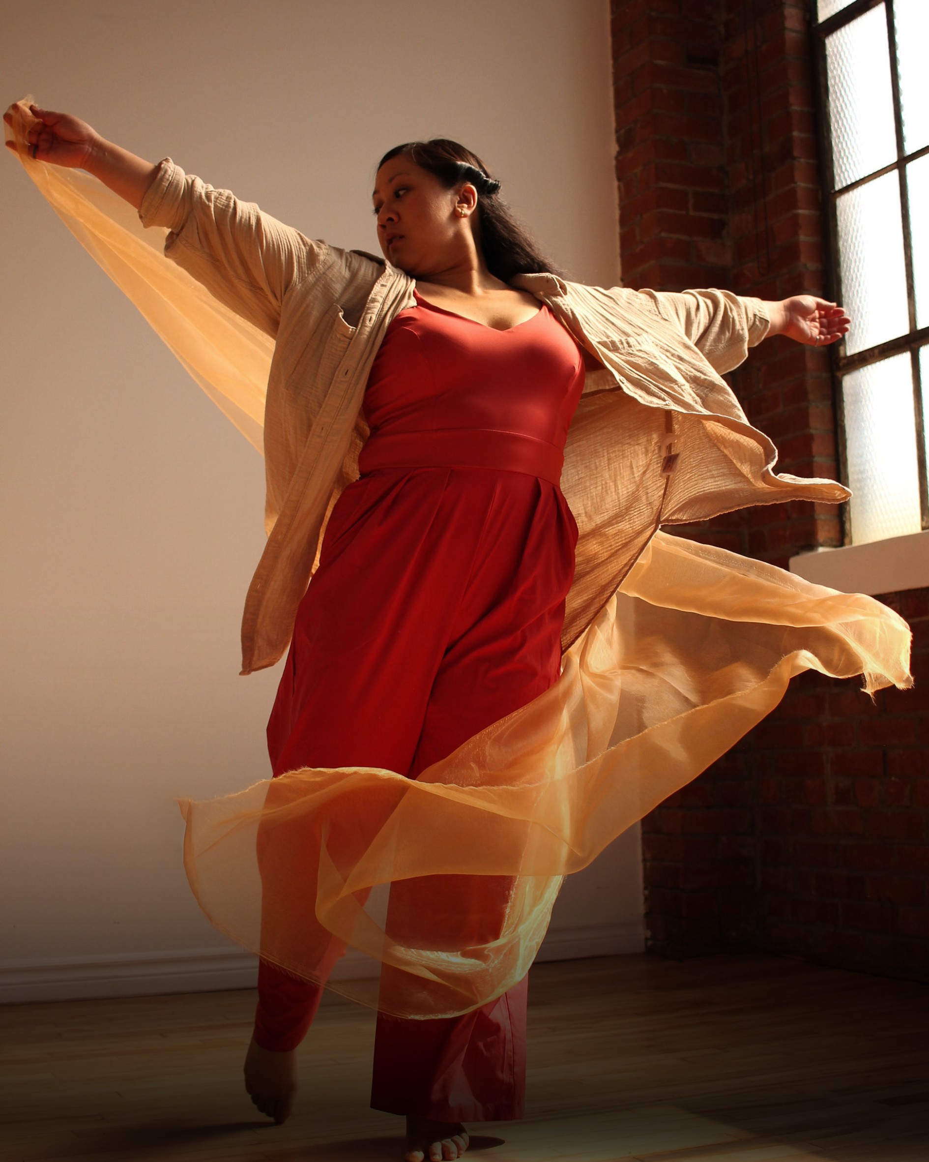 A woman dancing indoors near a large window with brick wall, wearing red pants and a tank top, with a beige shirt that flows as she moves.