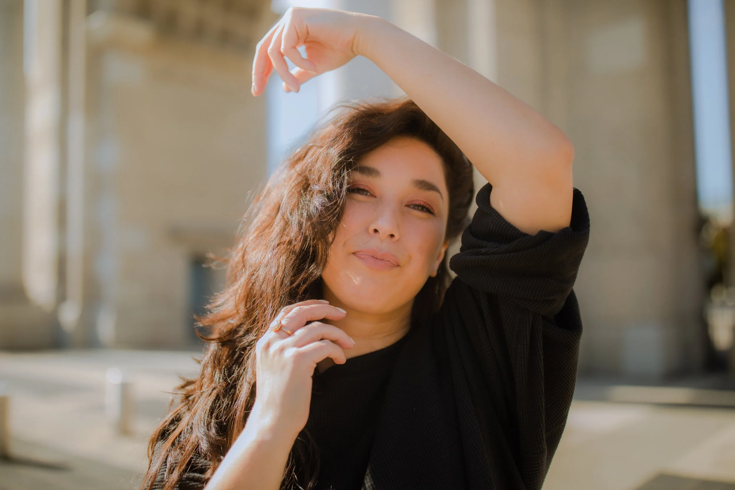 A woman with long brown hair, wearing a black top, poses outdoors with her arm over her head and a slight smile.