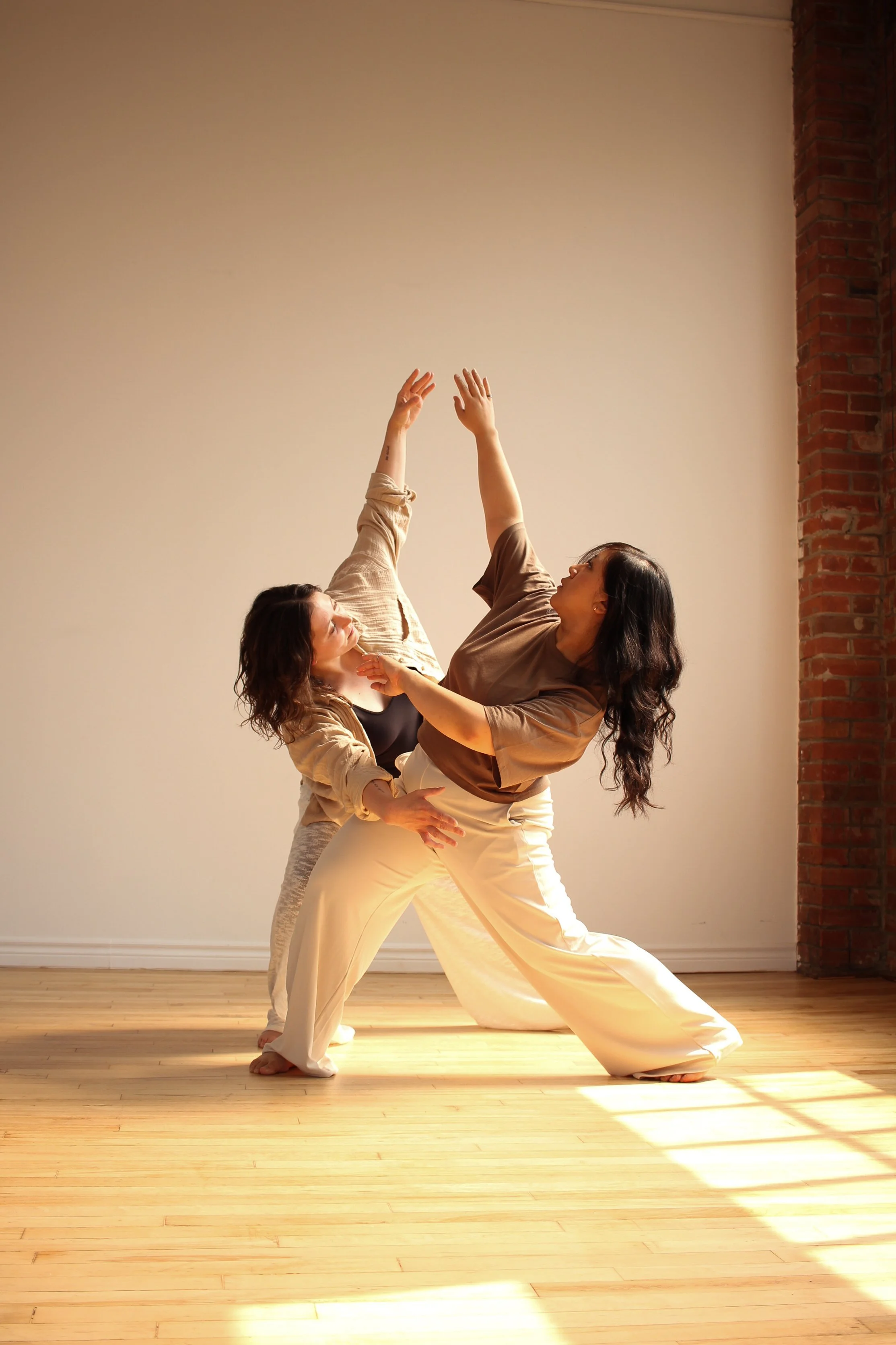 Two women practicing partner yoga or dance in a bright room with wooden flooring and a brick wall, performing a pose with one woman leaning back and the other supporting her.